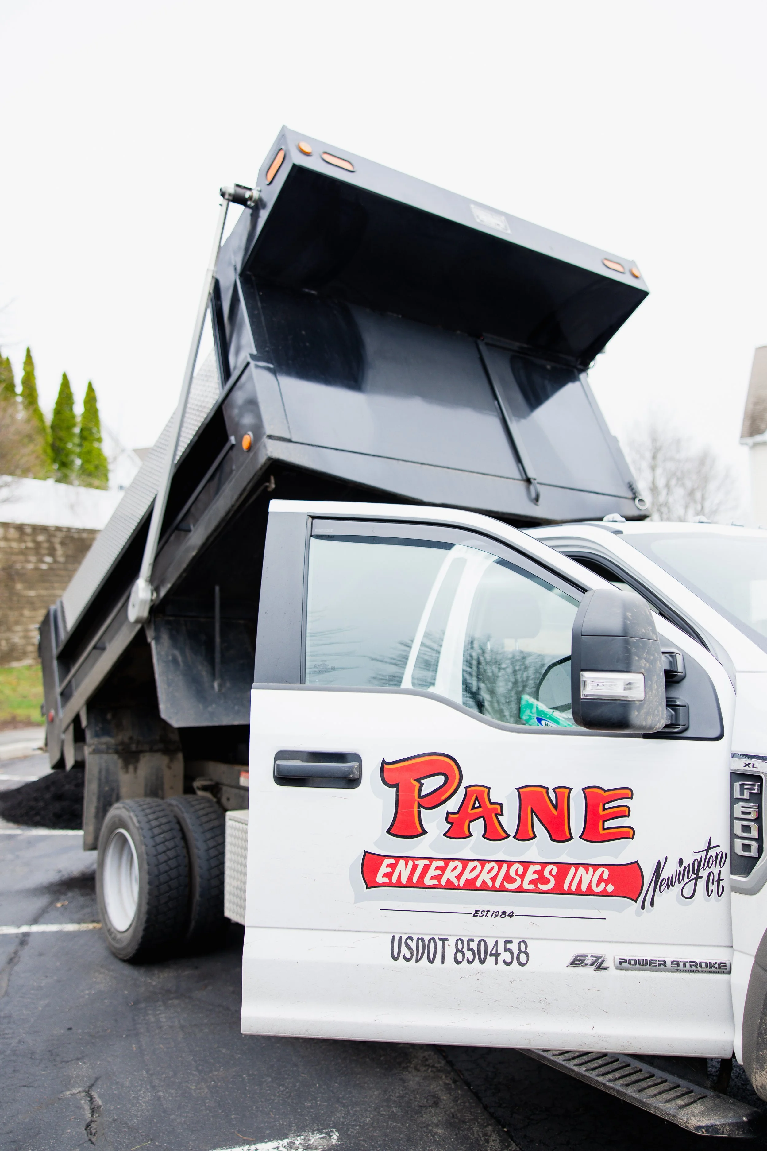 A white utility truck with a black elevated platform or lift on its bed. The truck has a business logo that reads "PANE ENTERPRISES INC." in large red and black letters. The location appears to be a parking lot with some trees and residential houses in the background on an overcast day.