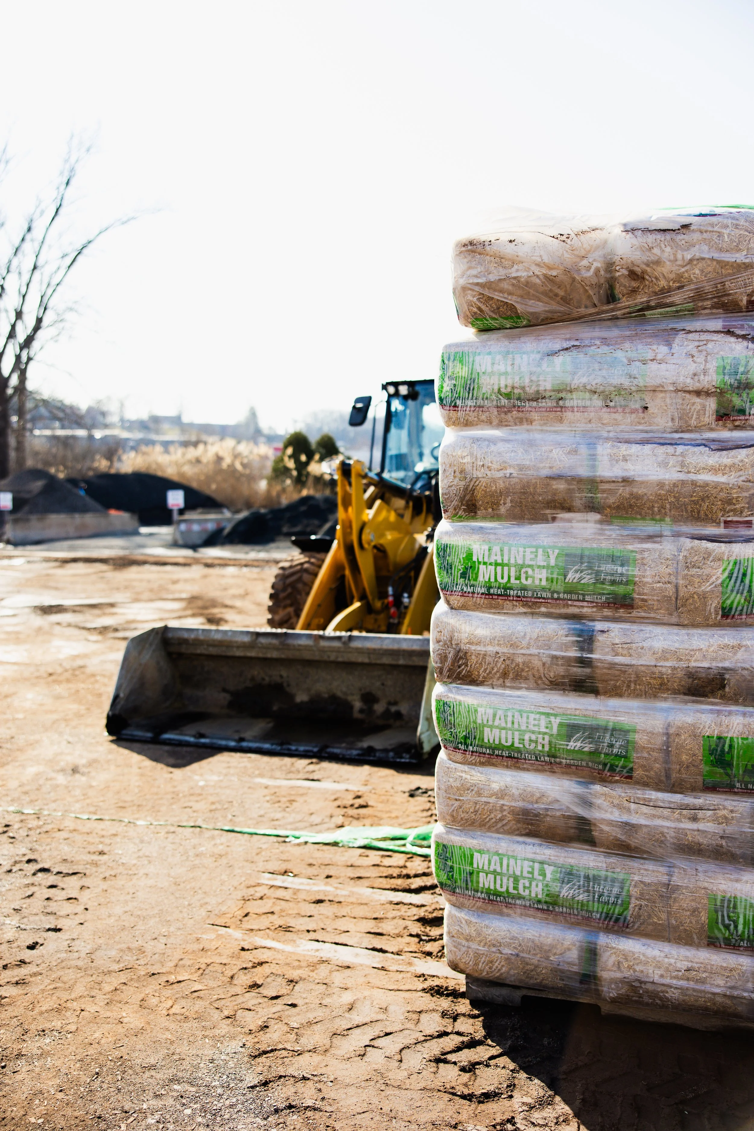 Stack of 4 pallets labeled 'Mainely Mulch' on a construction site with a bulldozer and piles of dirt in the background, overcast sky, no people present.