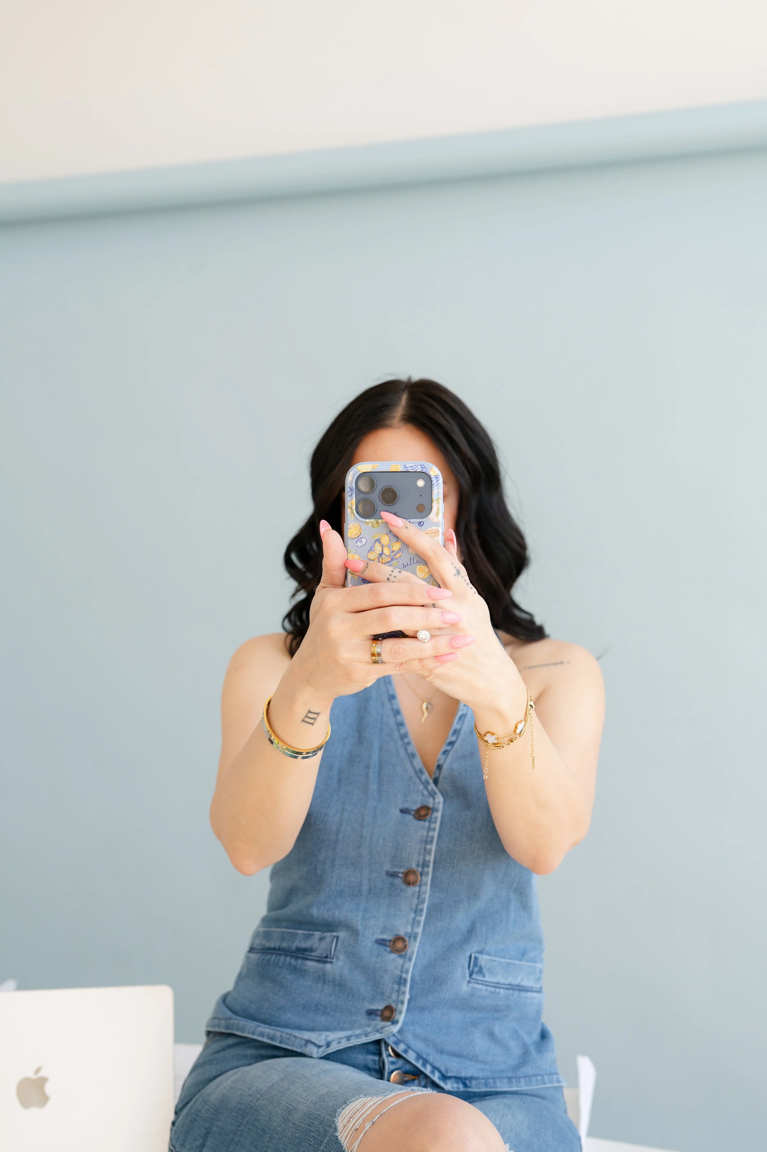 A woman taking a selfie with her phone, sitting in front of a light green wall, wearing a sleeveless denim dress, and a damaged ripped knee jeans, with jewelry on her arms and a tattoo under her right arm.