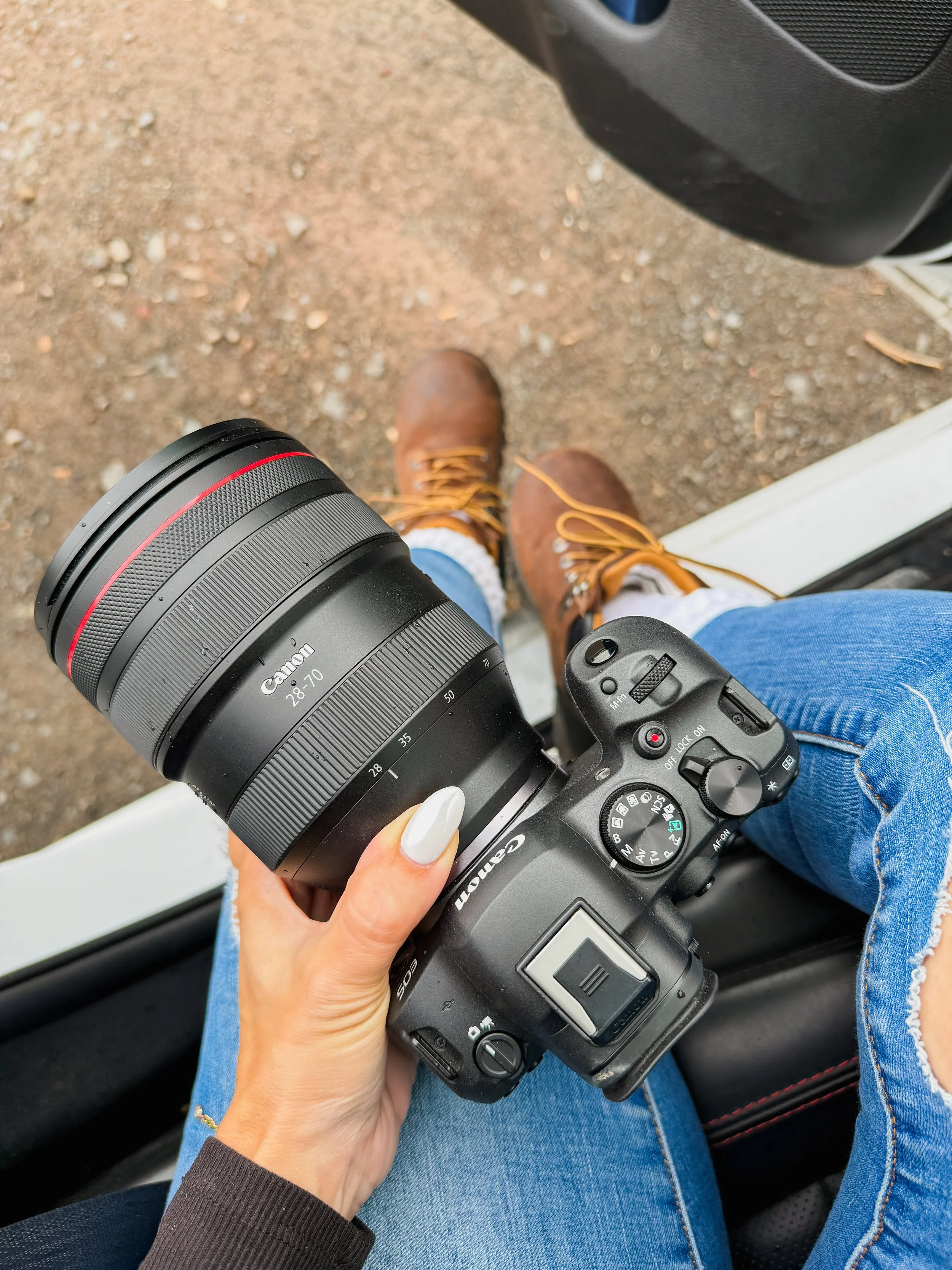 A person holding a Canon camera with a telephoto lens, sitting in a vehicle, wearing blue jeans and brown boots on a dirt ground outside.