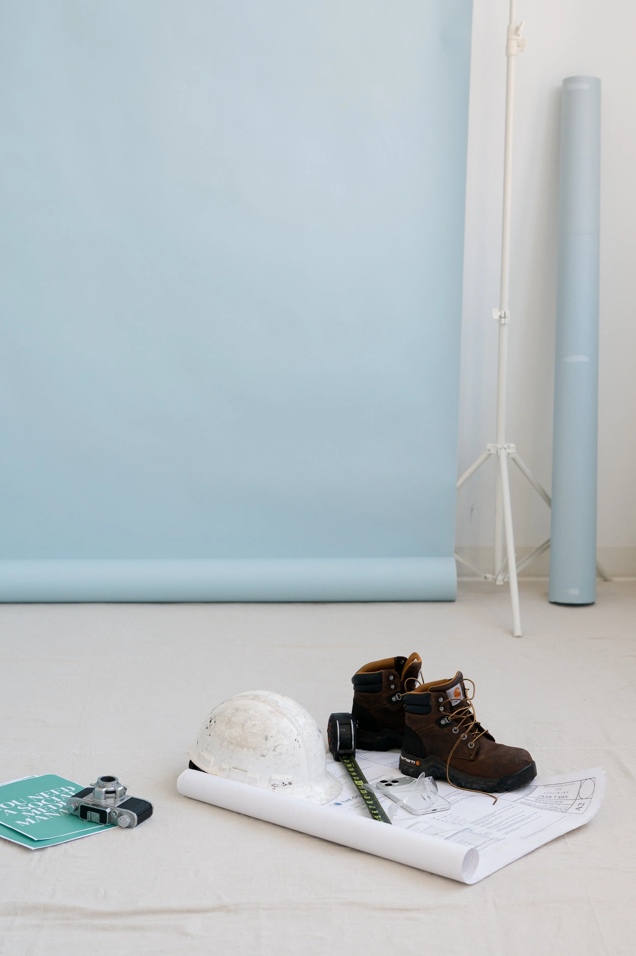 Construction workspace with a light blue backdrop, a pair of work boots, a white hard hat, a rolled-up blueprint, measuring tape, a small camera, and a pamphlet on safety.