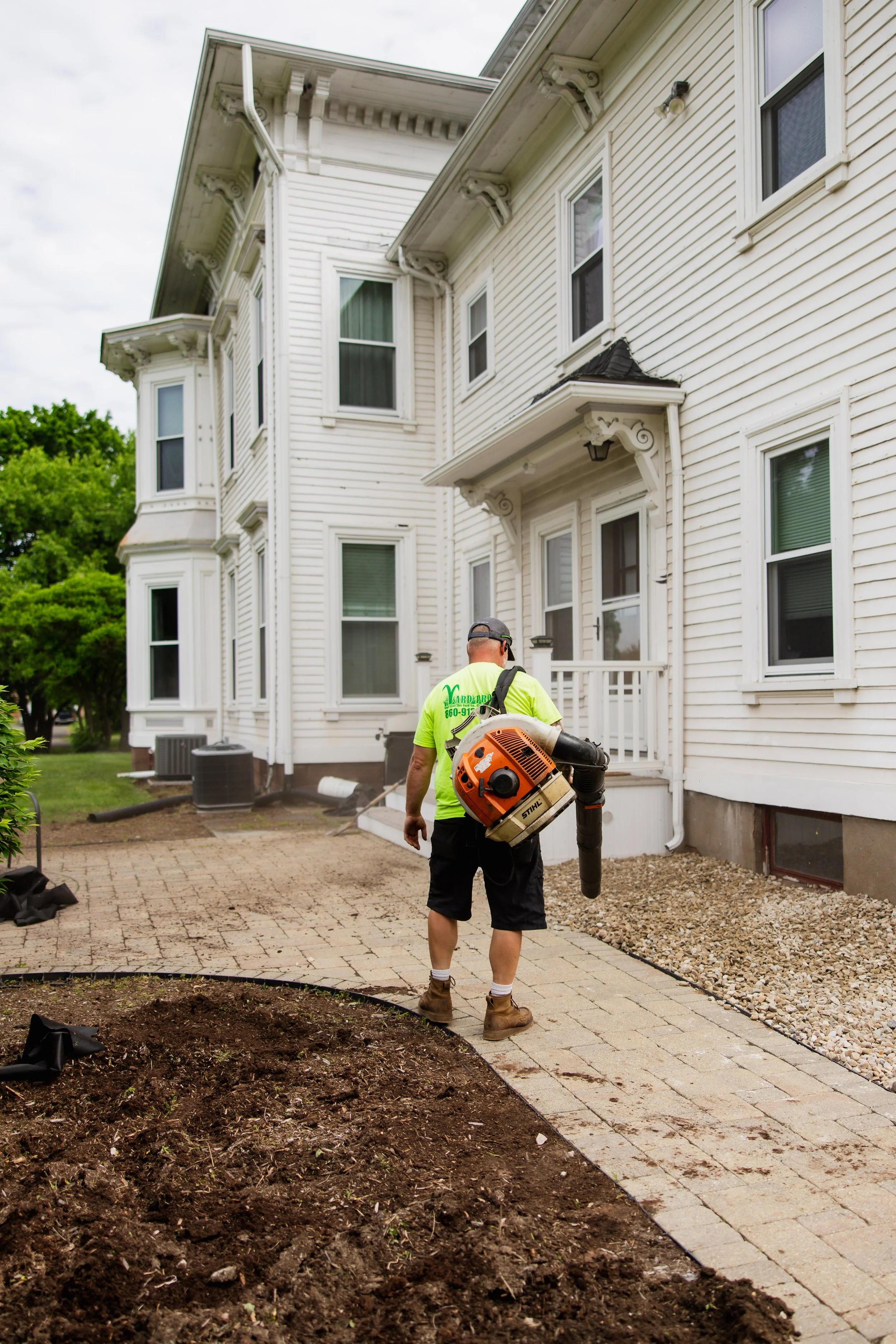 A worker wearing a green shirt, black shorts, and boots is using a leaf blower outside a white, three-story house with multiple windows and decorative architectural details.