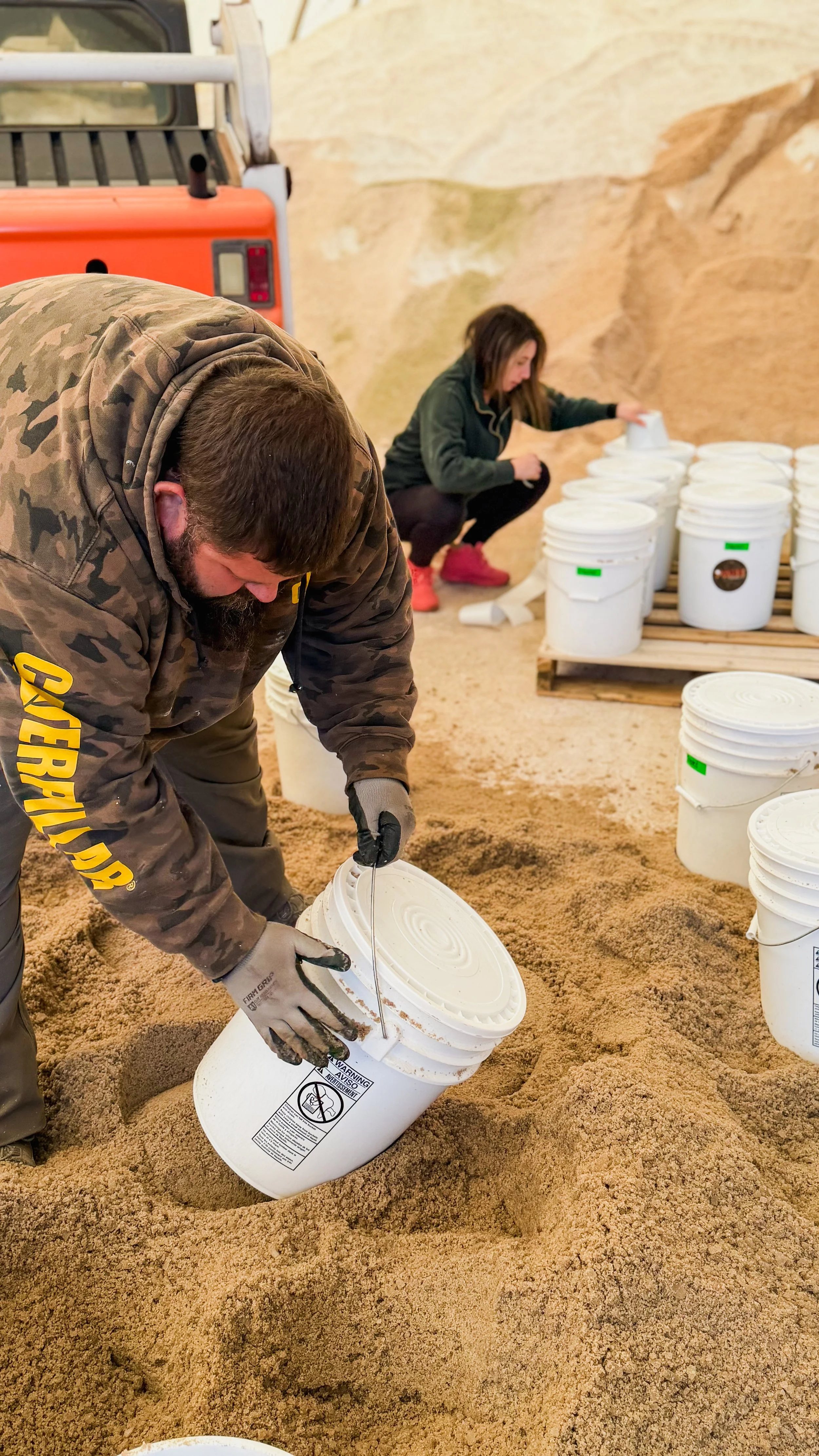 Two people are working with white buckets inside a sandy environment, possibly a cave or mine. The man in the foreground is wearing a camouflage jacket and gloves, pouring contents from a bucket. The woman in the background is crouching and handling 