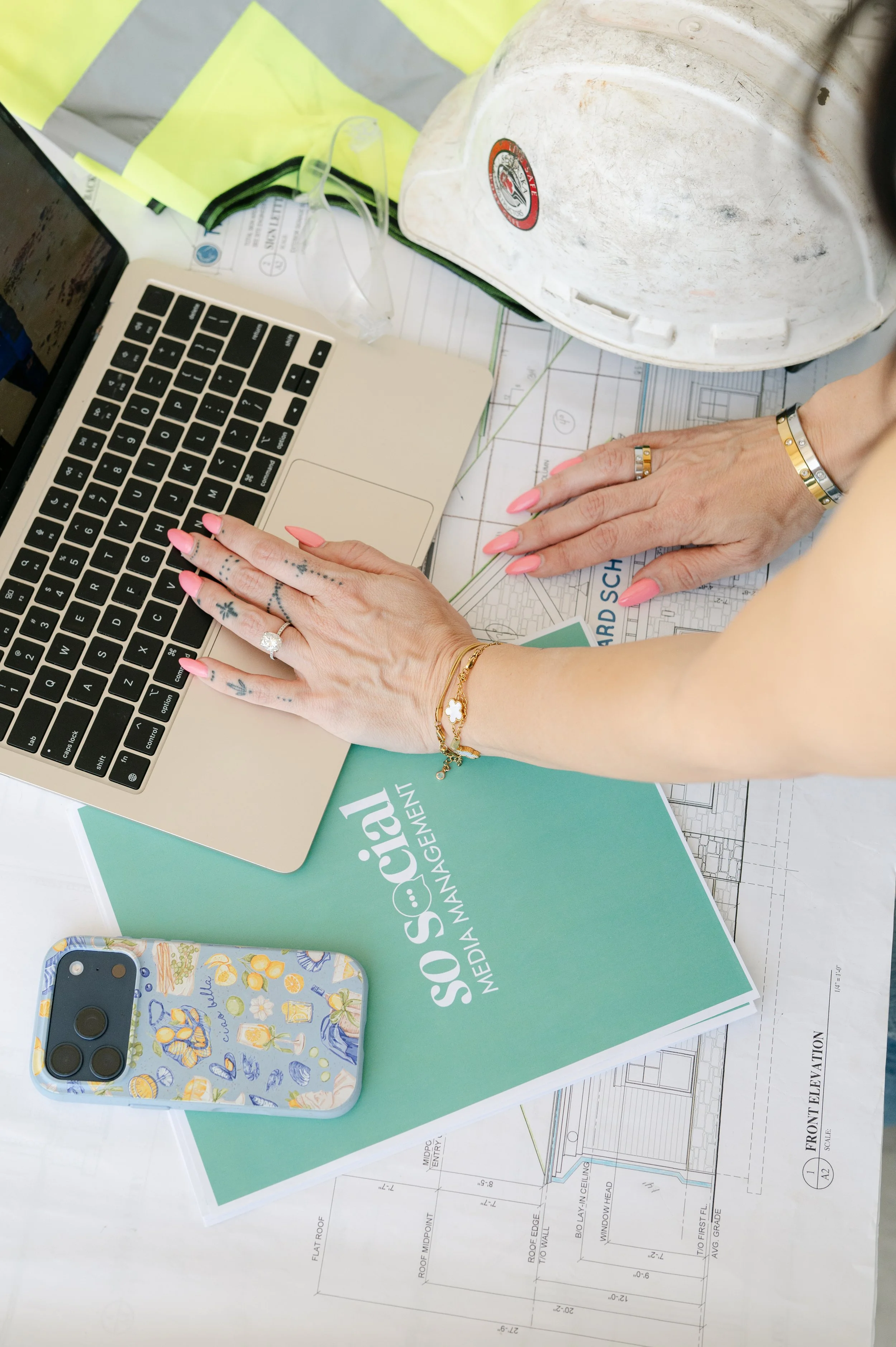 A person's hands working on architectural plans and a laptop on a desk, with construction helmet, safety glasses, and a smartphone nearby.