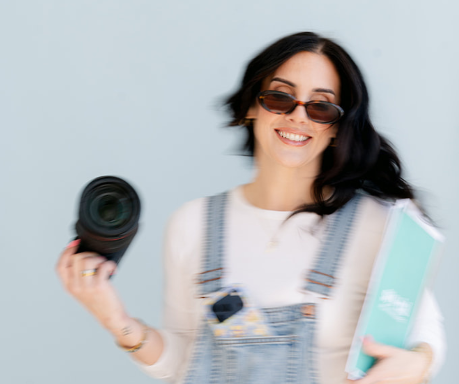 A woman with dark hair wearing sunglasses and overalls, smiling and holding a camera in one hand and a notebook in the other, standing against a light blue background.