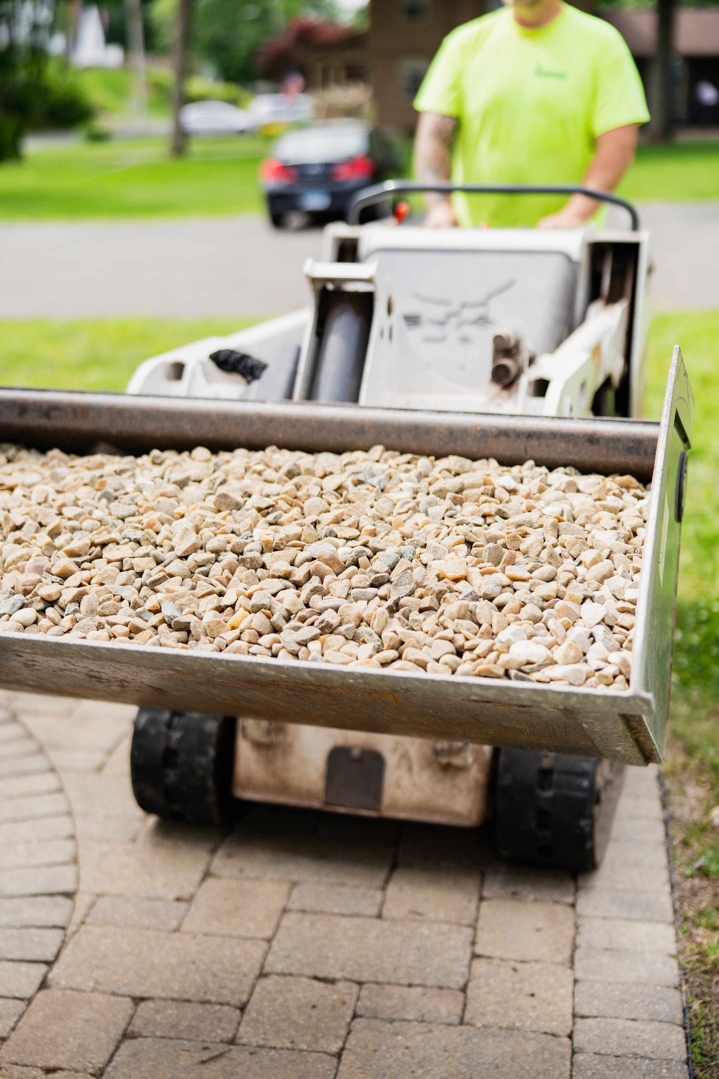 A person in a neon yellow shirt pushing a wheelbarrow loaded with small rocks on a brick patio with a background of grass, trees, and a house.
