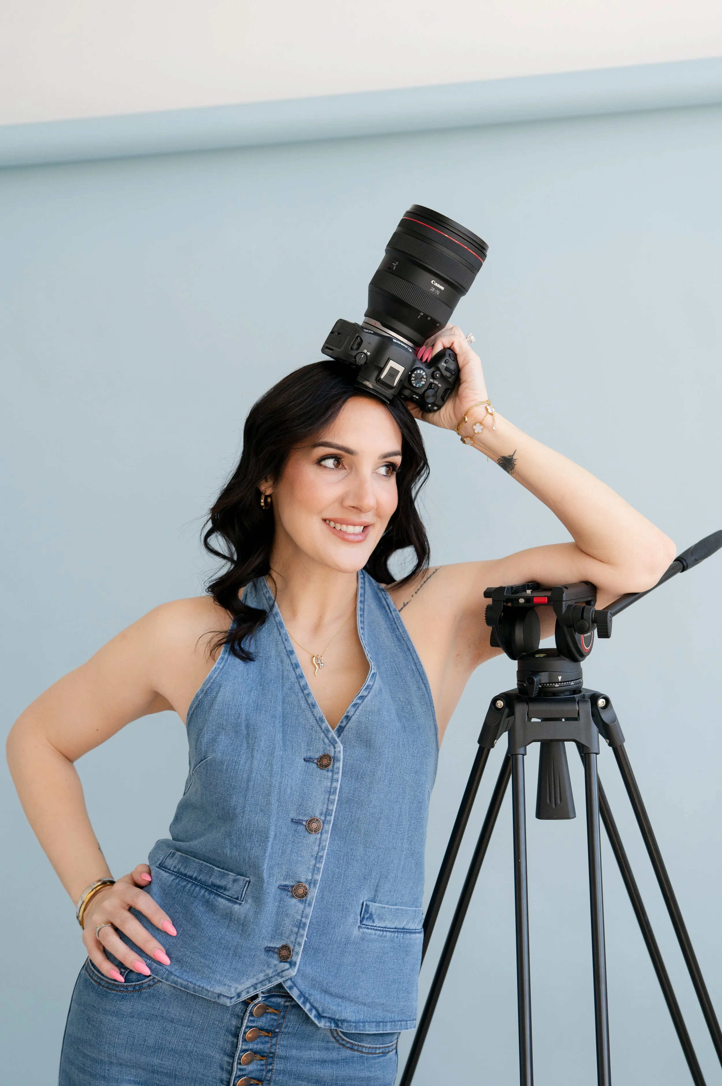 A woman with dark hair wearing a sleeveless denim top, standing next to a camera tripod, holding a professional camera with a large zoom lens on her shoulder, smiling and posing with one hand on her hip against a light blue wall.