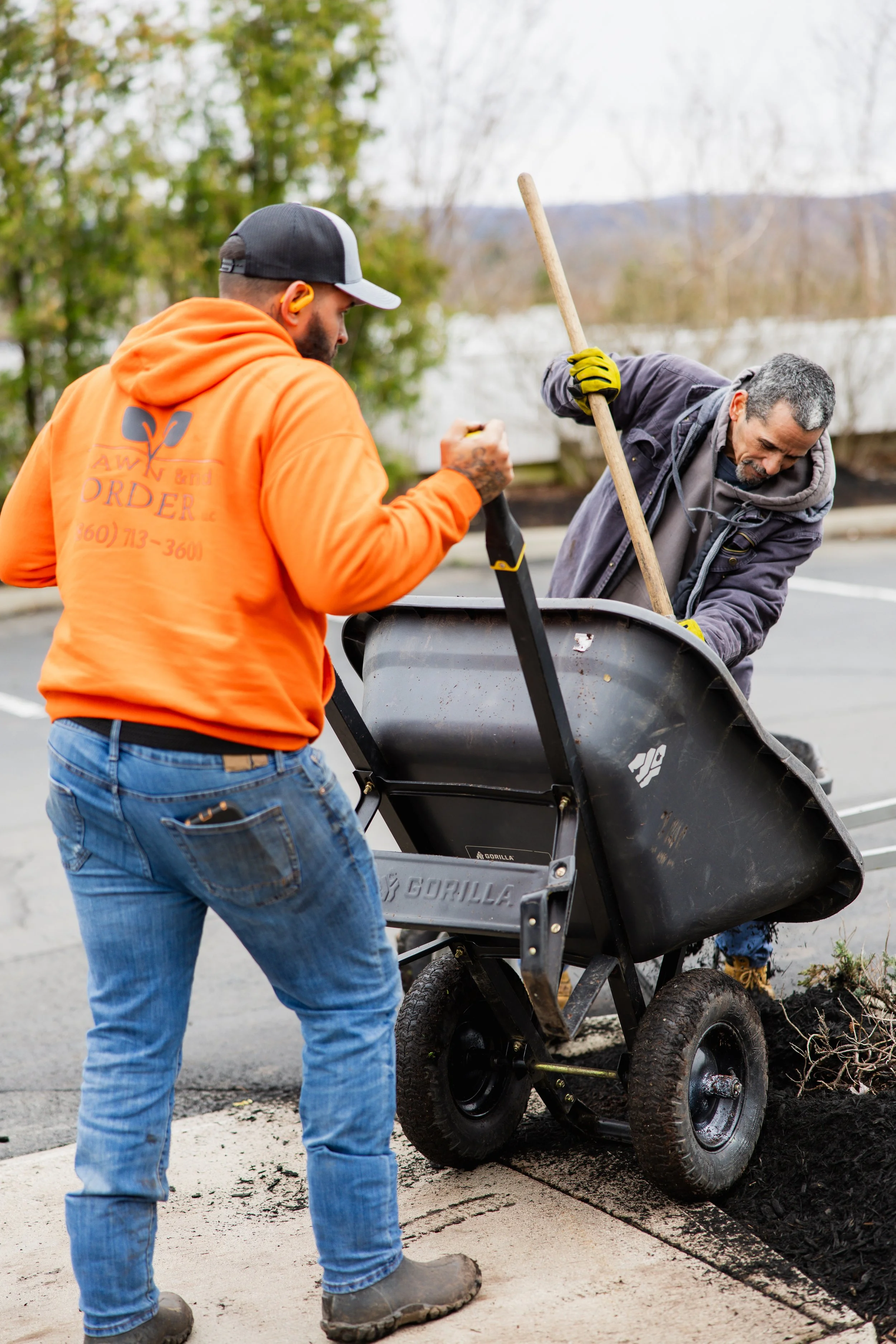 Two men working outdoors near a street; one man in an orange hoodie and jeans operates a wheelbarrow filled with dirt or debris, while the other man in a gray jacket and jeans leans into the wheelbarrow, using a tool; trees and a cloudy sky are in th