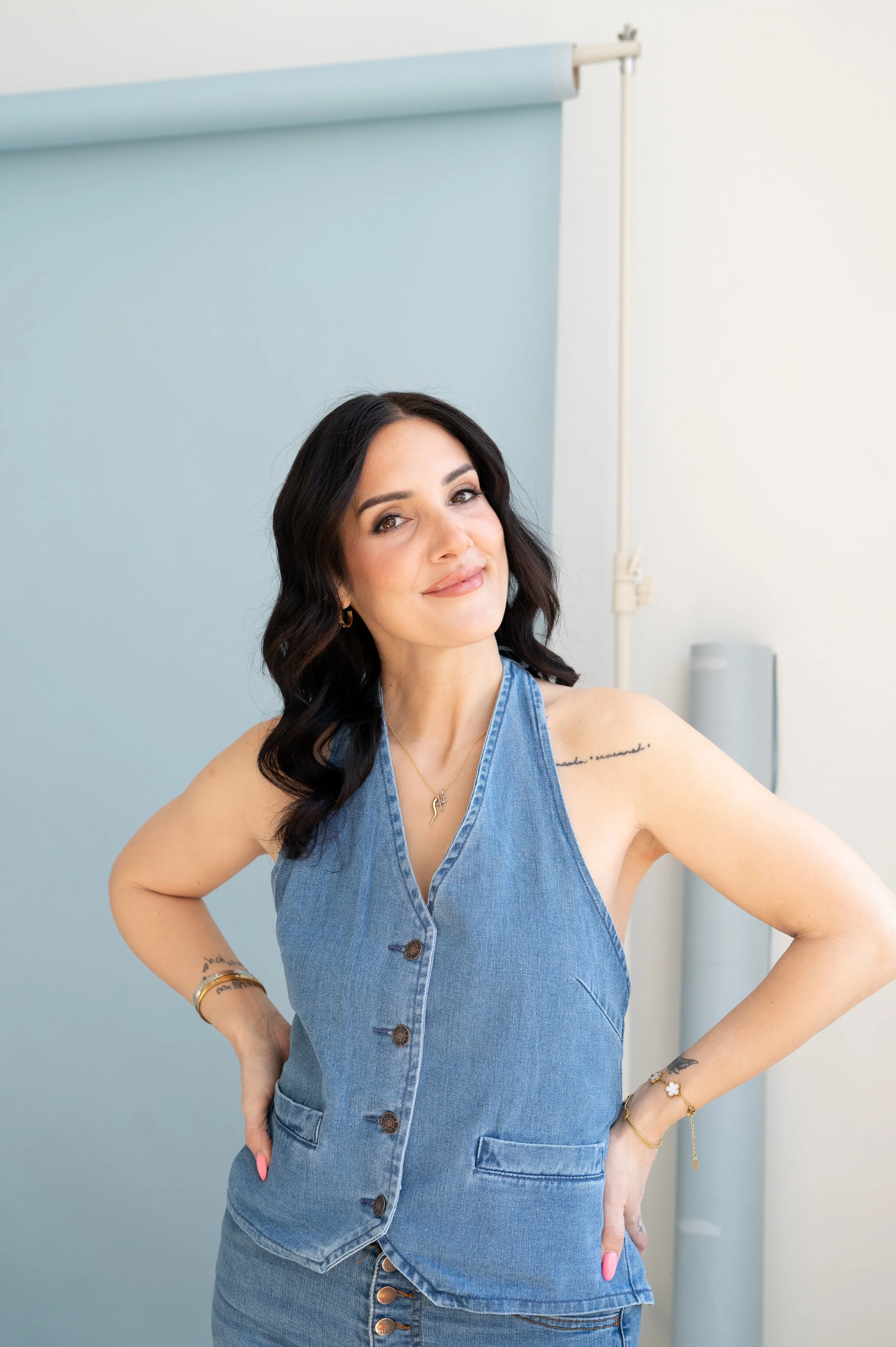 A woman with long dark hair wearing a sleeveless denim dress, standing with one hand on her hip, smiling slightly, in front of a light blue backdrop in a photo studio.