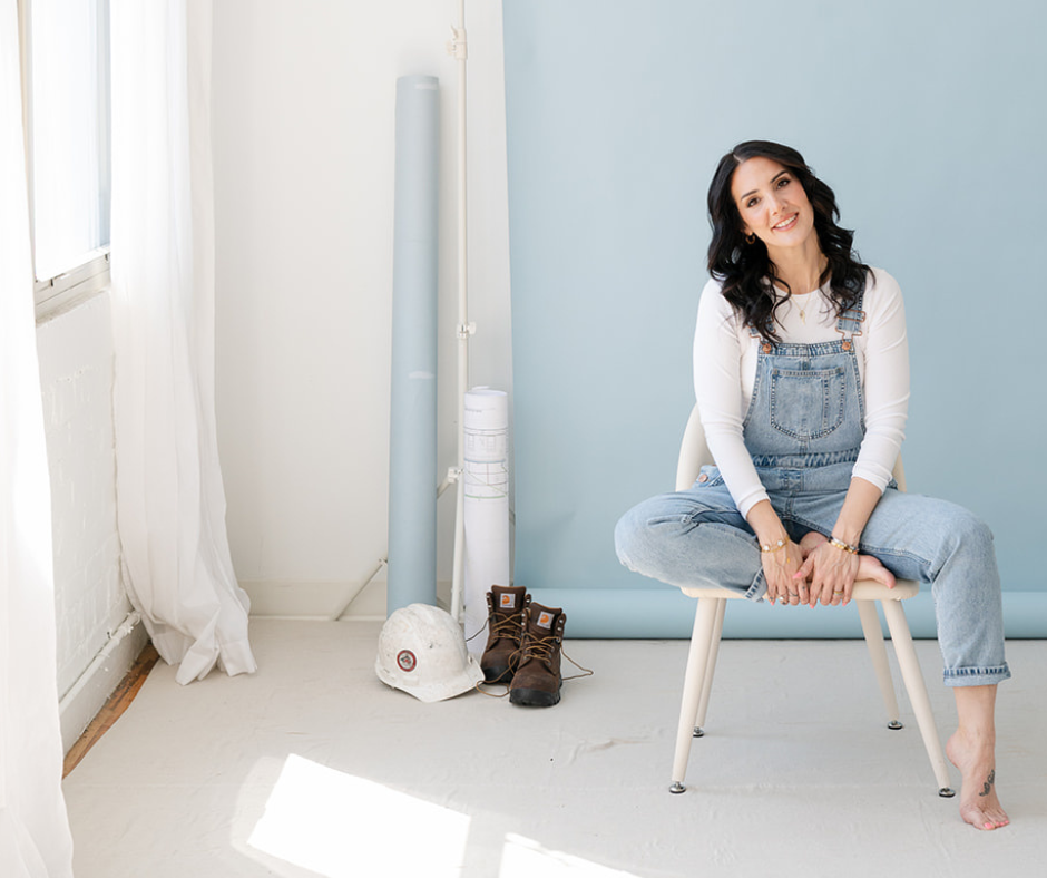 A woman with dark hair wearing overalls and a white shirt sitting on a chair in a bright studio, with construction materials and gear around her.
