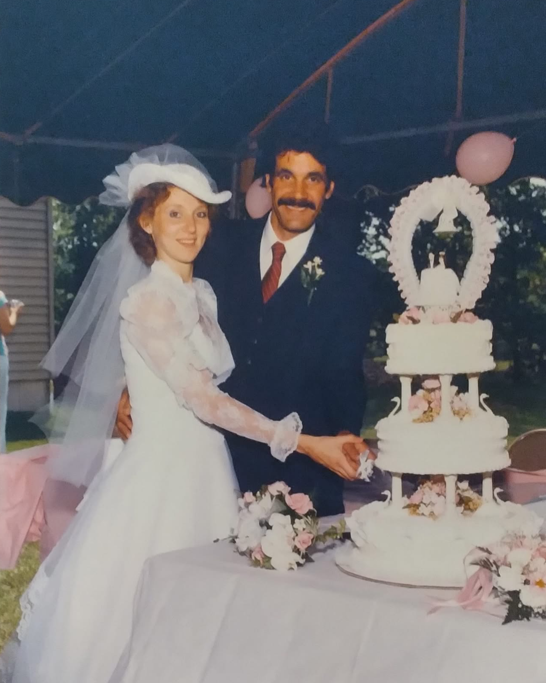 A bride and groom at their wedding cake, smiling and holding hands, outdoors under a canopy decorated with pink balloons, on a table decorated with flowers.