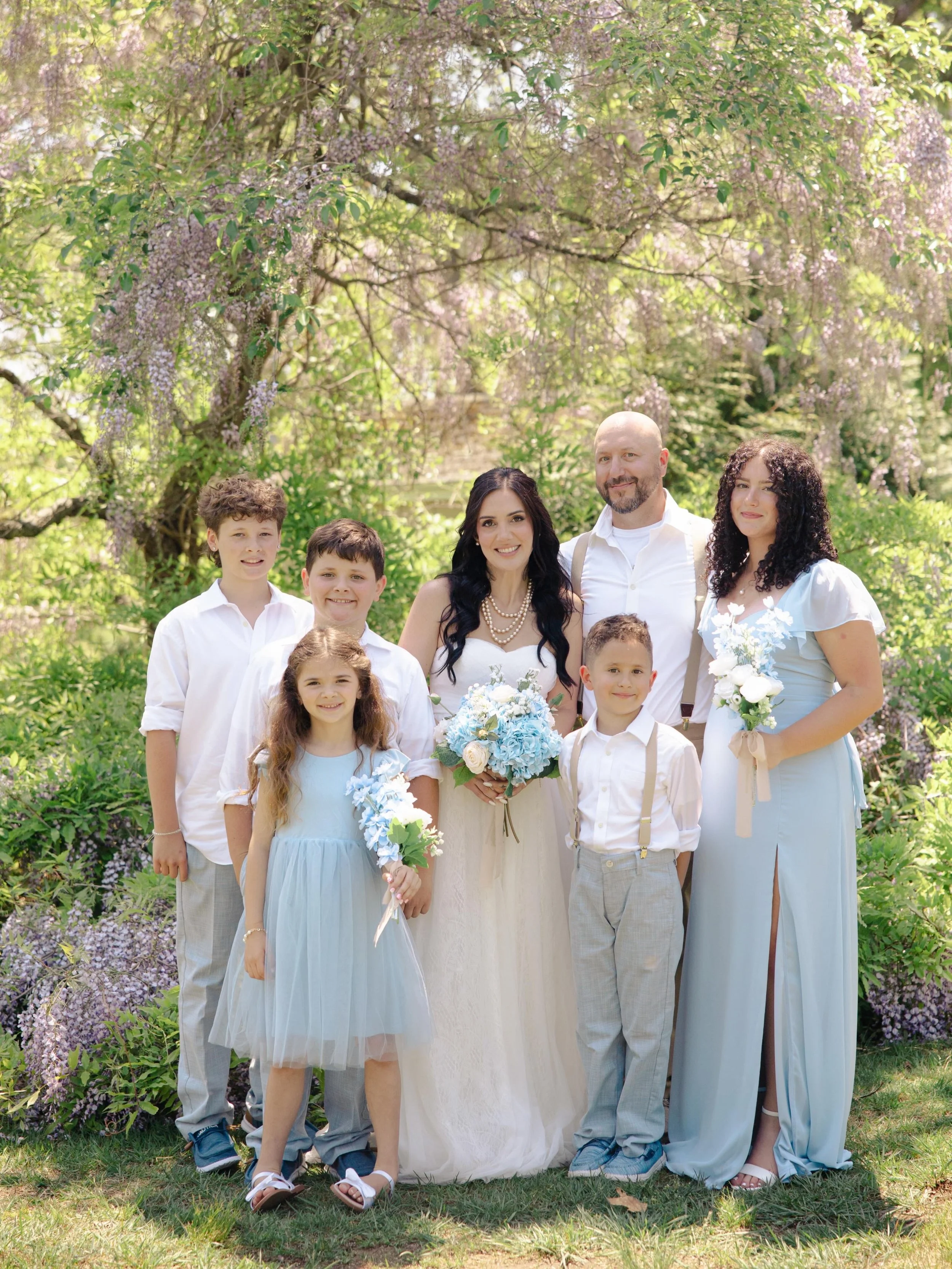 A family of seven standing outdoors in front of pink flowering trees, dressed in light-colored spring clothing, some holding bouquets.