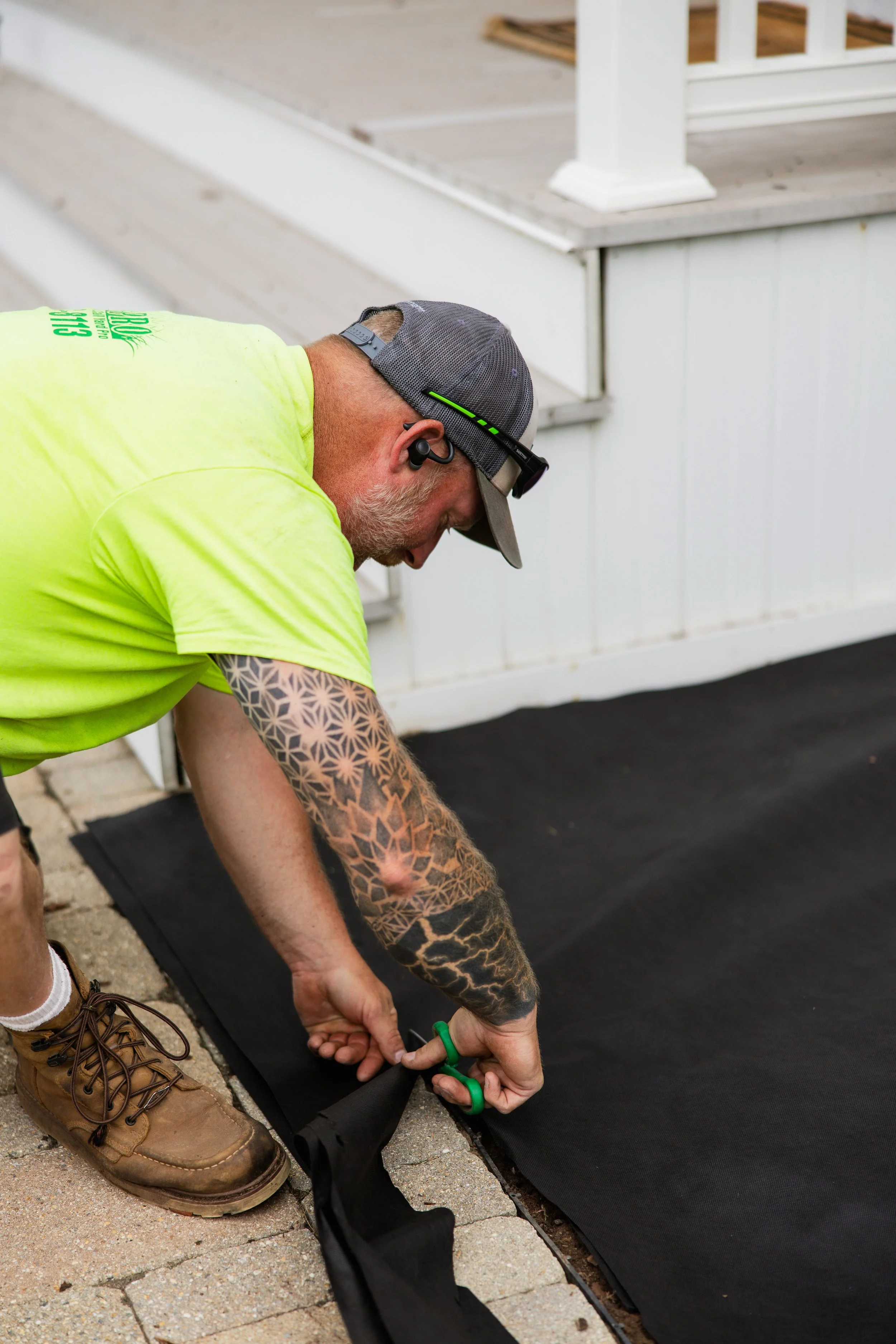 A man with tattoos on his arms wearing a gray cap, yellow shirt, and brown work boots is kneeling on a brick patio, using green scissors to roll out a black fabric or tarp on the ground. There is a white porch in the background.