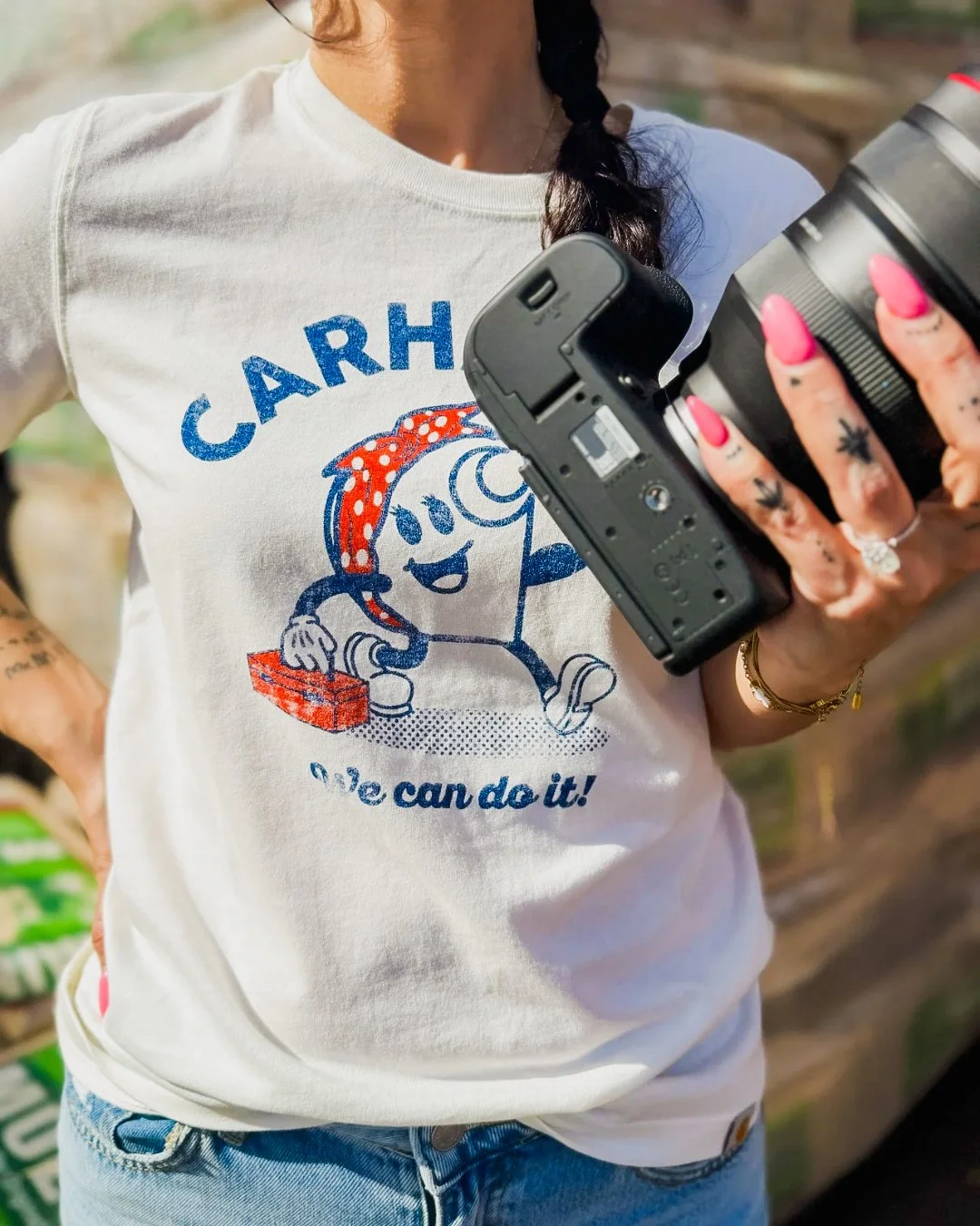 Person wearing a white t-shirt with a cartoon character and the text 'CARH! We can do it!' holding a camera, with visible tattoos and pink nail polish.