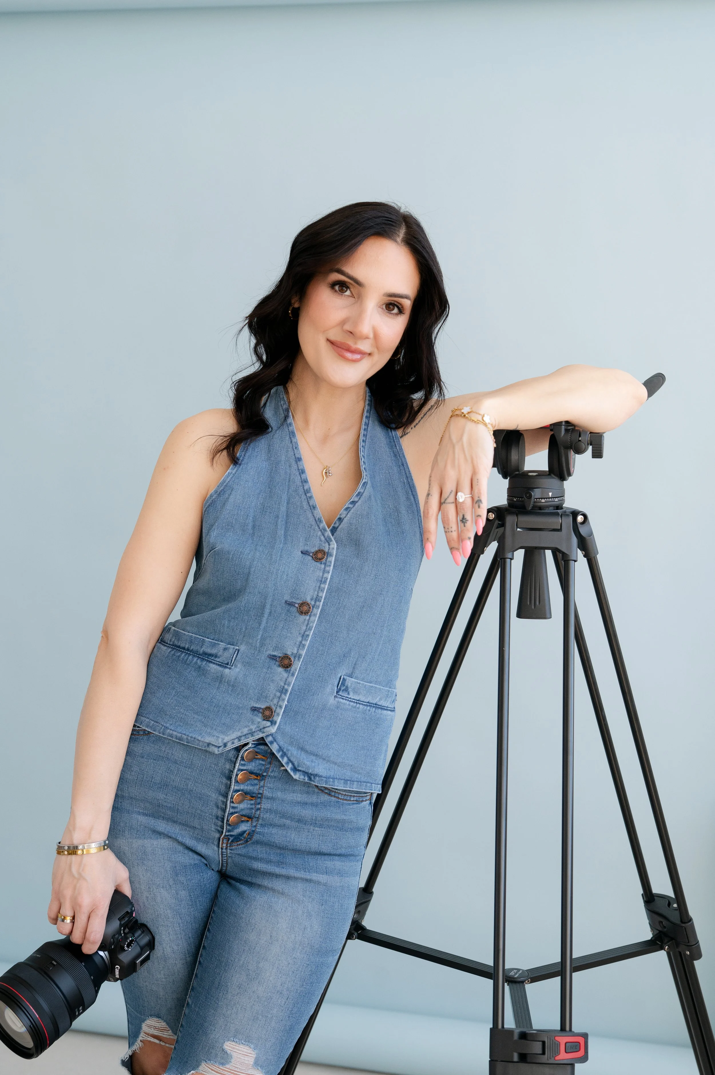 A woman in a denim vest and jeans holding a camera in her right hand, posing beside a camera tripod, with a light blue background.