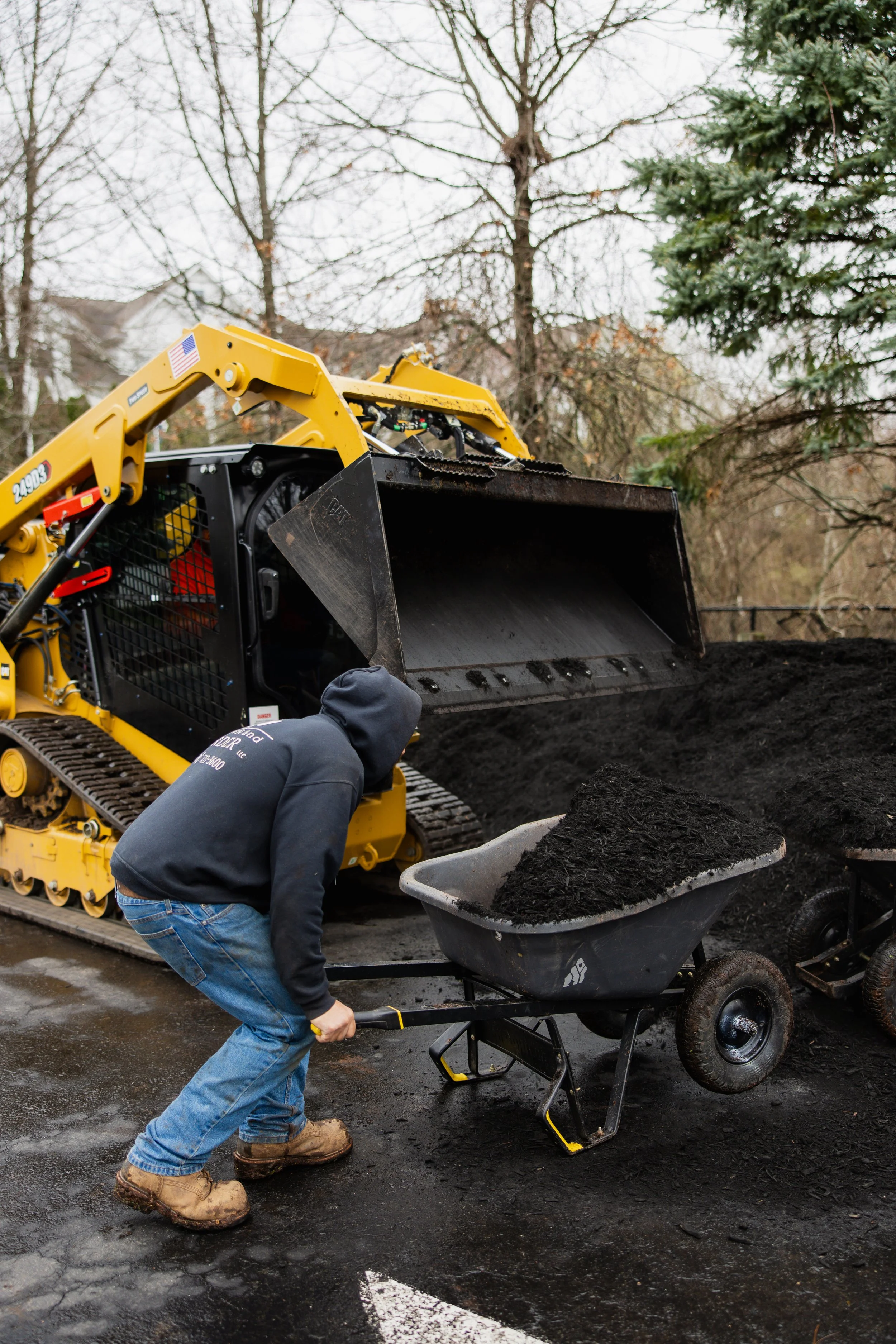 A worker in blue jeans, a grey hoodie, and tan boots is loading dark soil from a wheelbarrow into a black machine. In the background, a yellow construction machine is visible along with trees and houses.