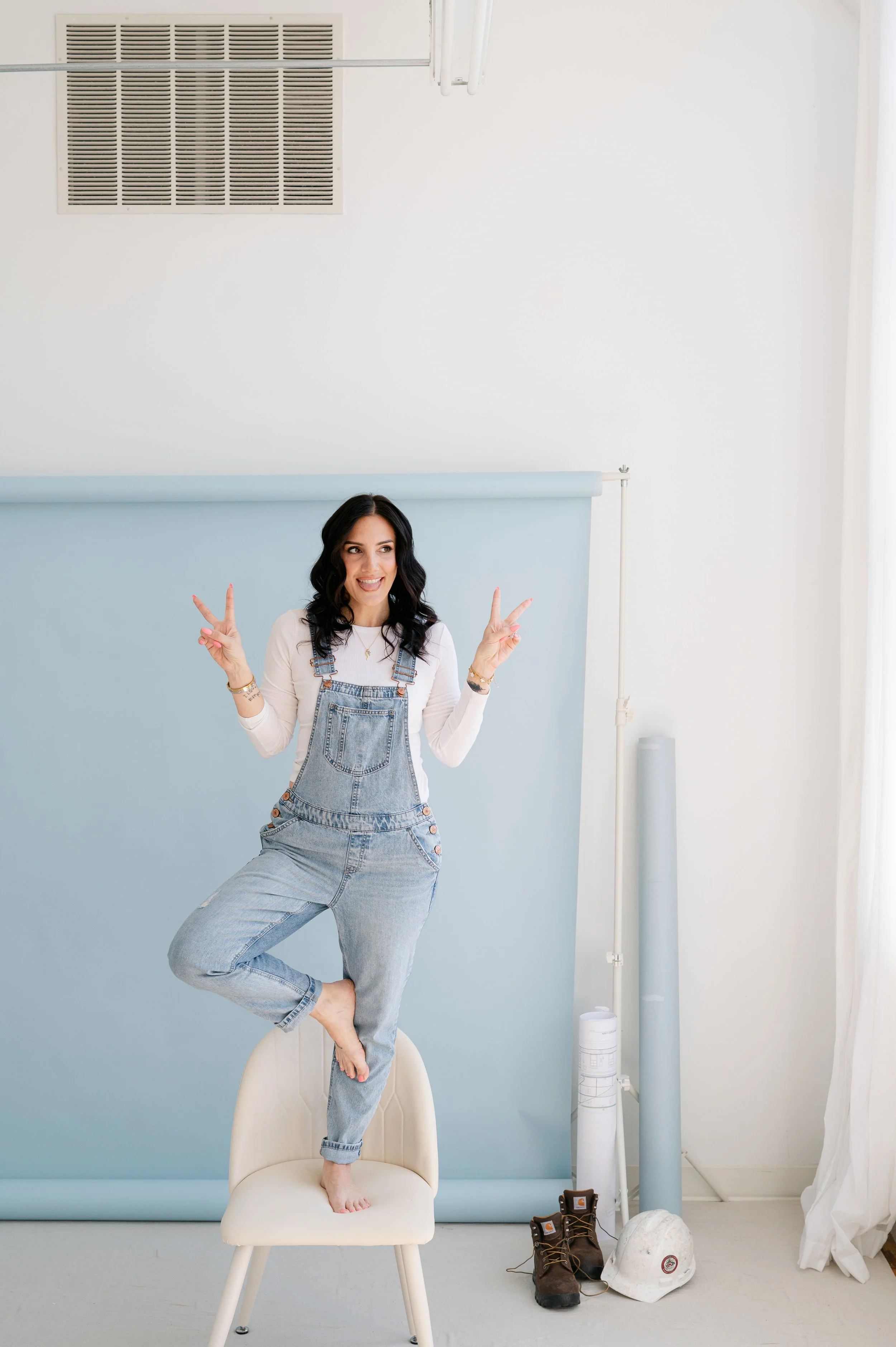 A woman with black hair wearing a white long sleeve shirt and denim overalls is balancing on one leg on a chair, posing with peace signs near her face, smiling in a room with construction tools and a light blue wall.