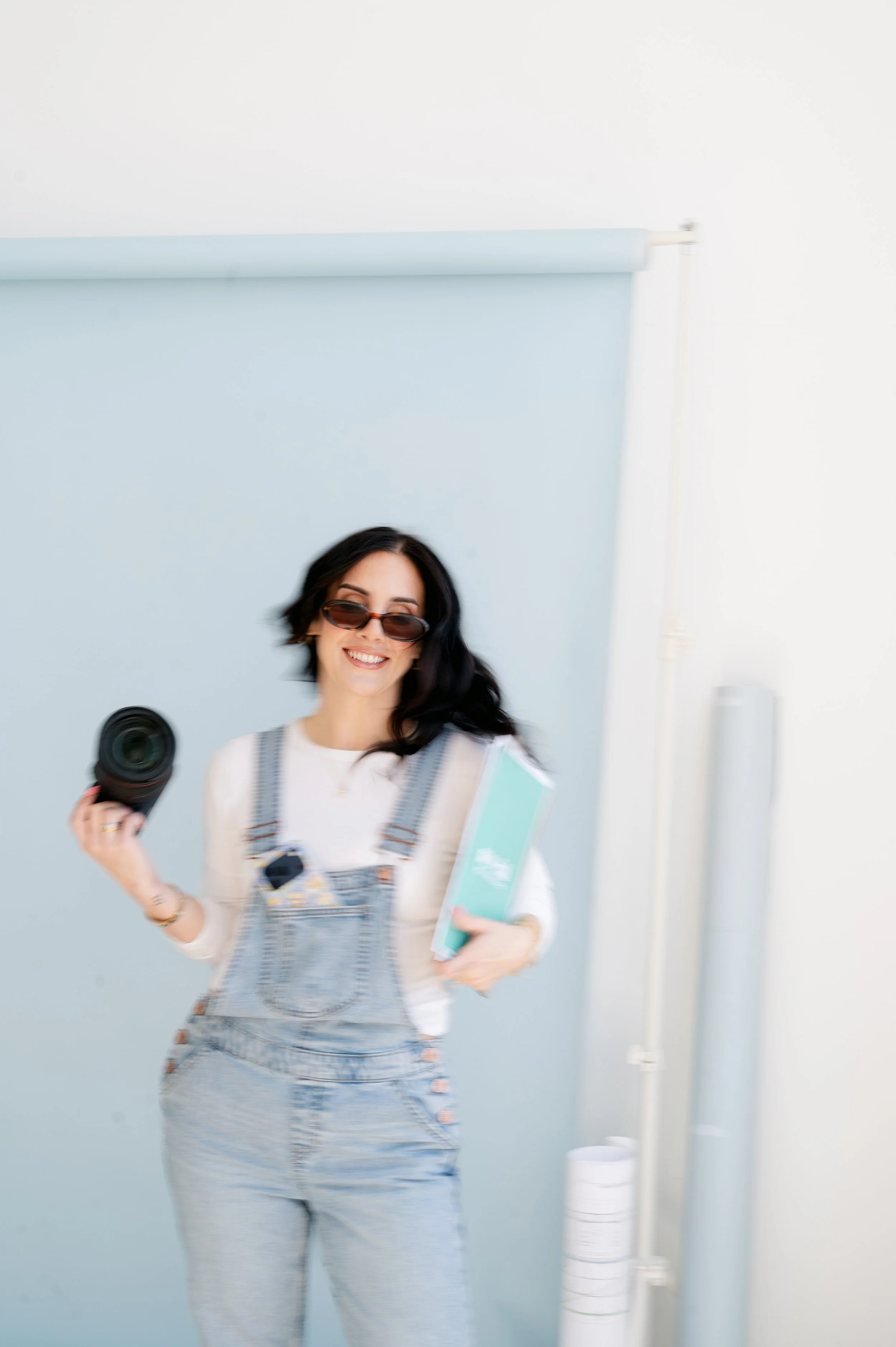 A woman with dark hair, wearing sunglasses, smiling, holding a camera in one hand and a book or folder in the other, standing in front of a light blue backdrop.