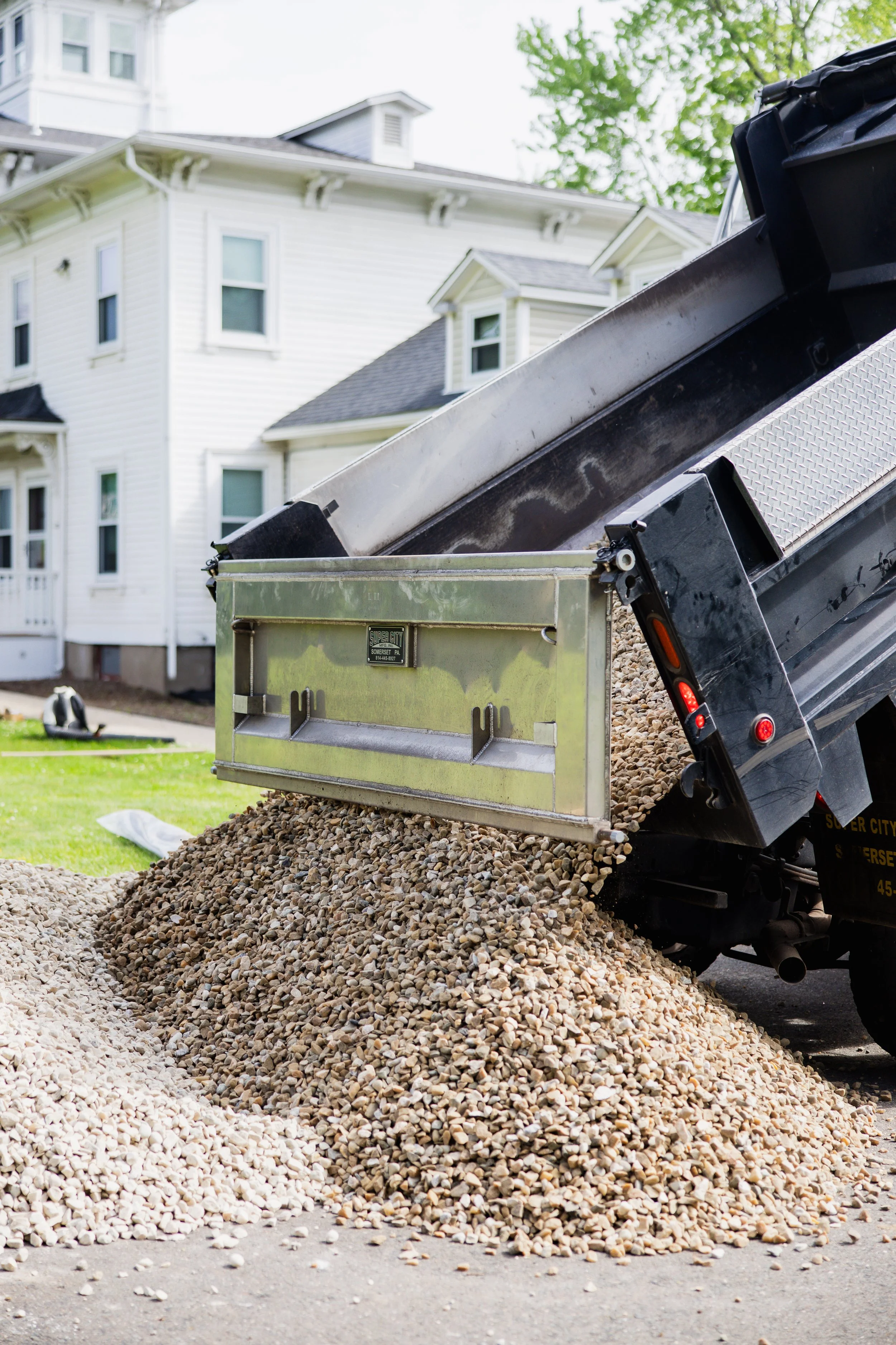A dump truck unloading a pile of small rocks or gravel on a driveway in front of a white house with multiple windows.