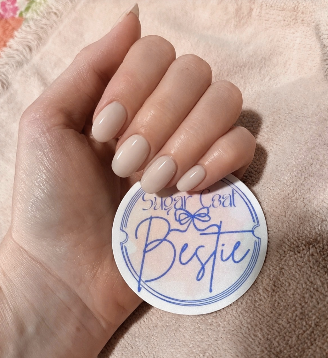 A hand with neatly manicured, glossy, beige-colored nails resting on a soft pink towel. The hand holds a round sticker that reads 'Sugar Cat Bustie' in decorative blue cursive and print fonts with a small bow illustration.