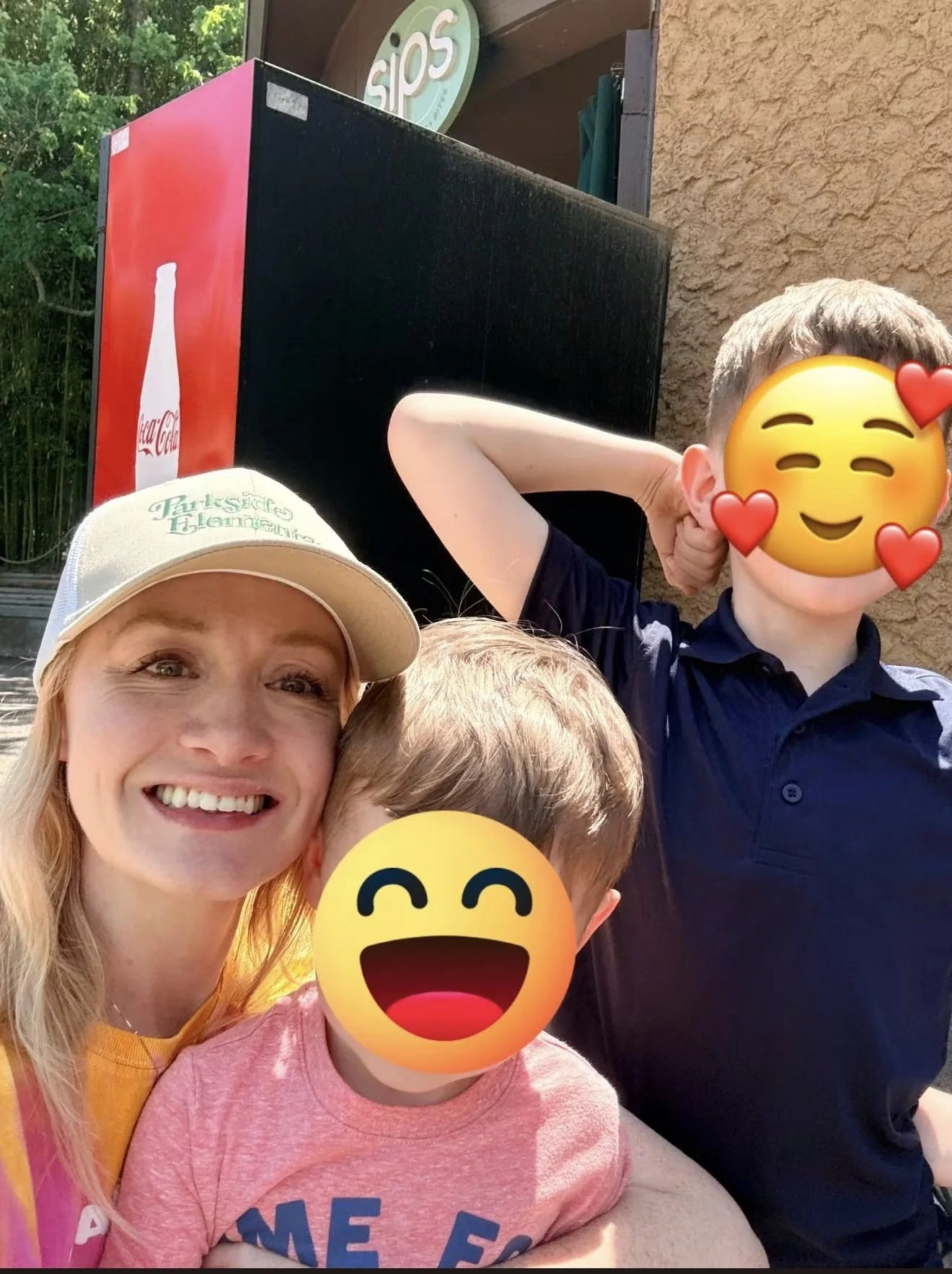 Group of four people taking a selfie outside on a sunny day, with tree leaves and a building in the background. Their faces are partially covered with emoji stickers, and they are smiling and showing relaxed, cheerful expressions.