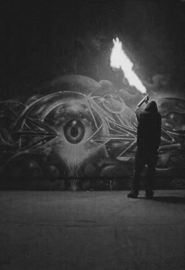 VYAL, seen from behind, spraying a mural of a large surreal eye at night