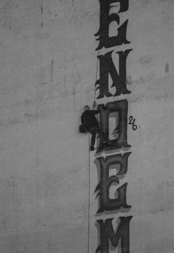 A graffiti writer on a rope rappelling beside large ENDEM letters painted down the side of a building