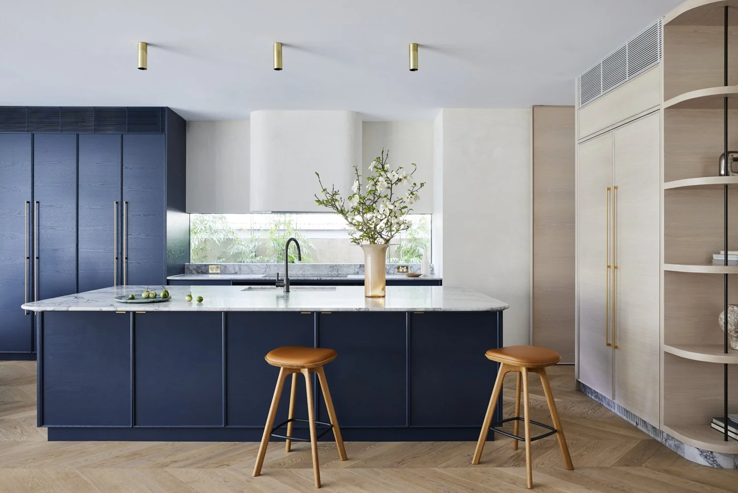 Modern kitchen with dark blue cabinets, a white marble island, a large flower vase, and wooden bar stools.