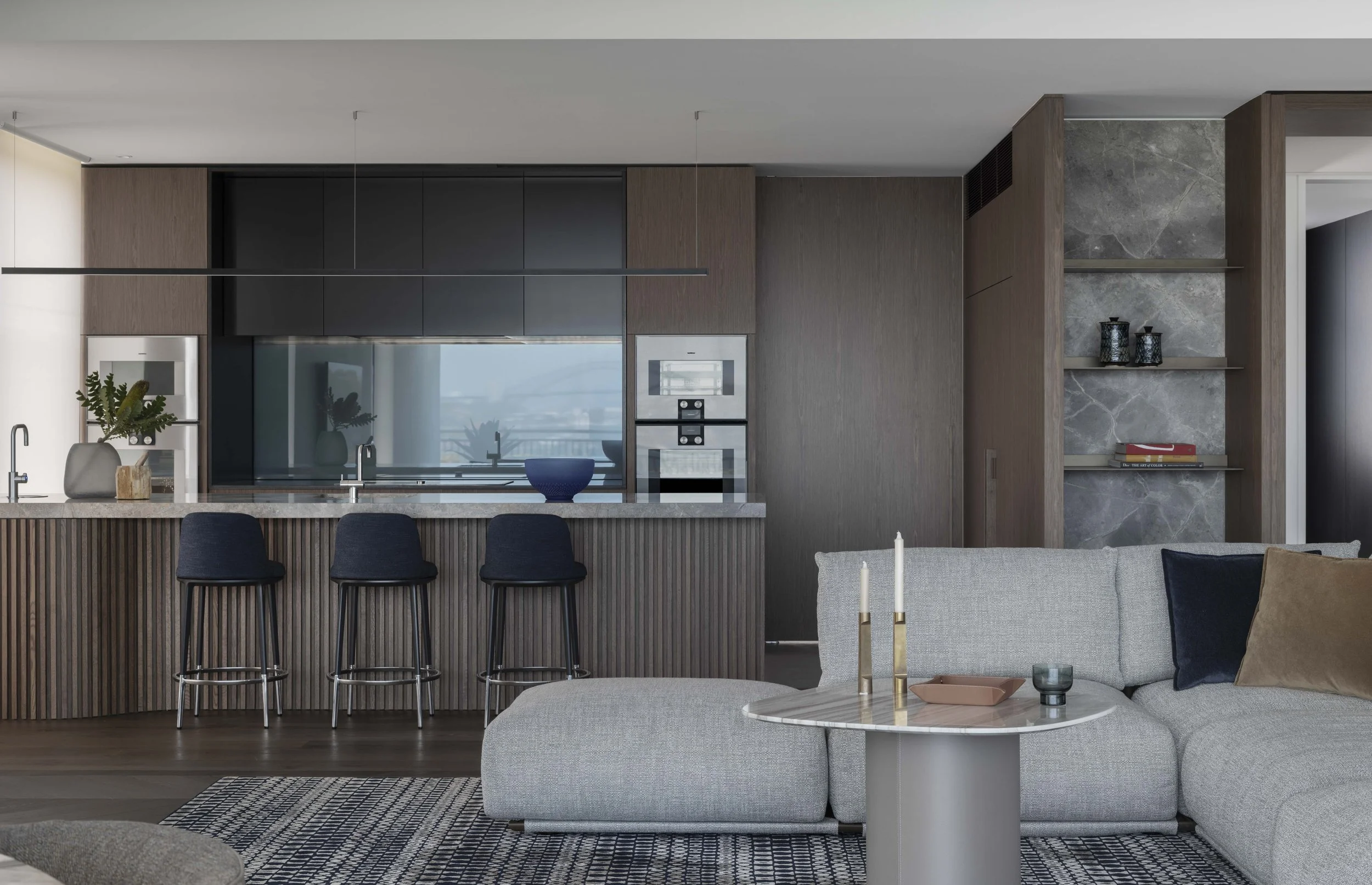 Modern open-plan kitchen and living room with grey sofa, marble, wood, and black accents, kitchen island with bar stools, decorative vases, and a view of a cityscape through a large window.