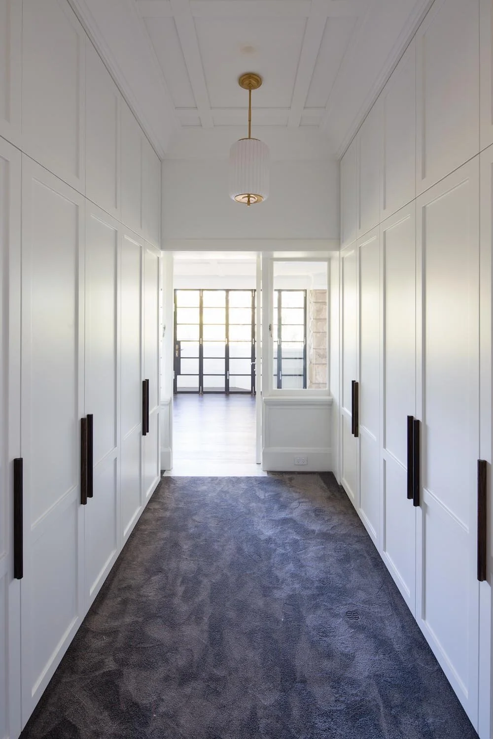 A hallway with white paneled walls, a dark carpeted floor, a ceiling light fixture, and glass doors at the end letting in natural light.