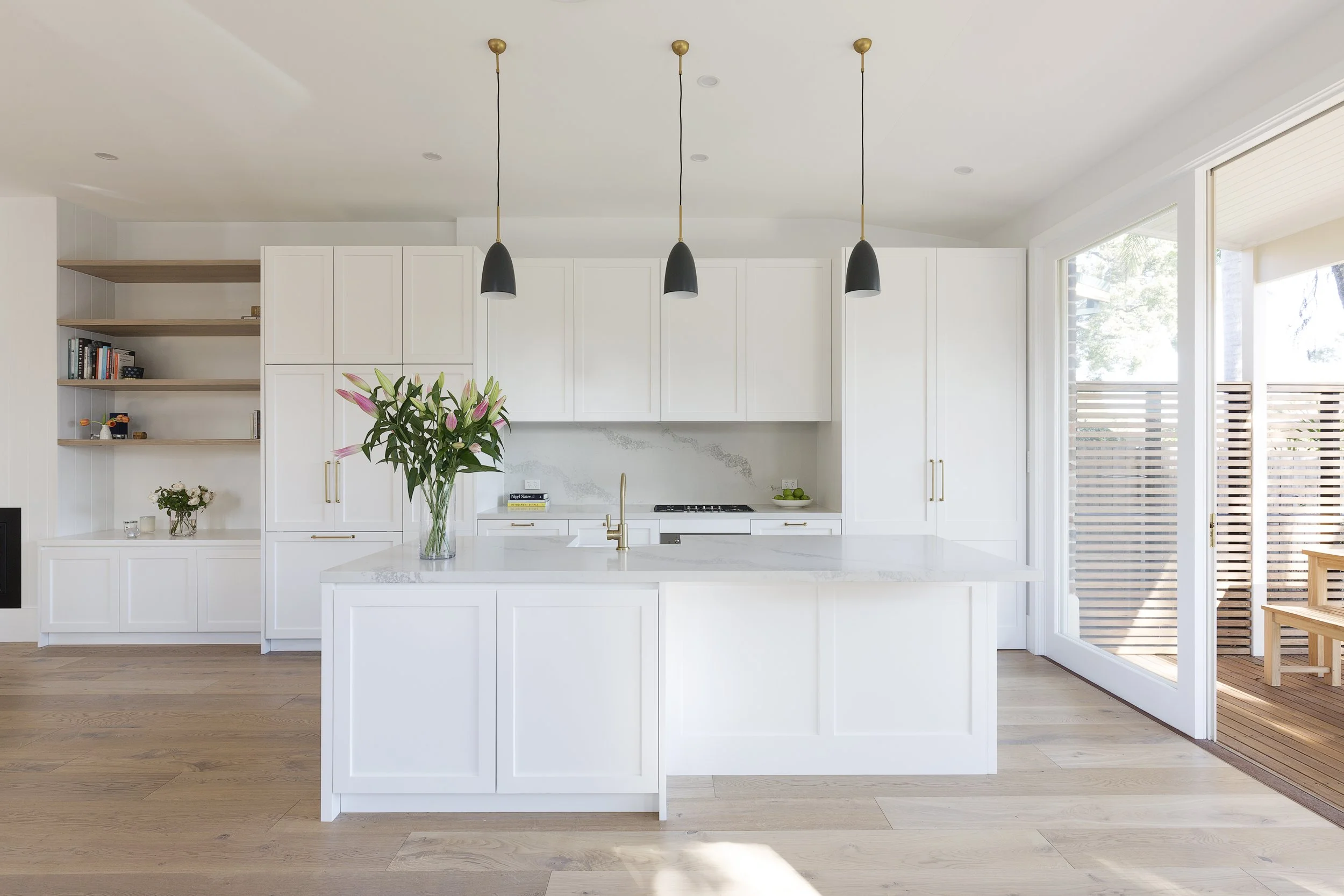 Modern white kitchen with a large island, black pendant lights, a stack of books, a vase with pink lilies, and sliding glass doors opening to a wooden deck.