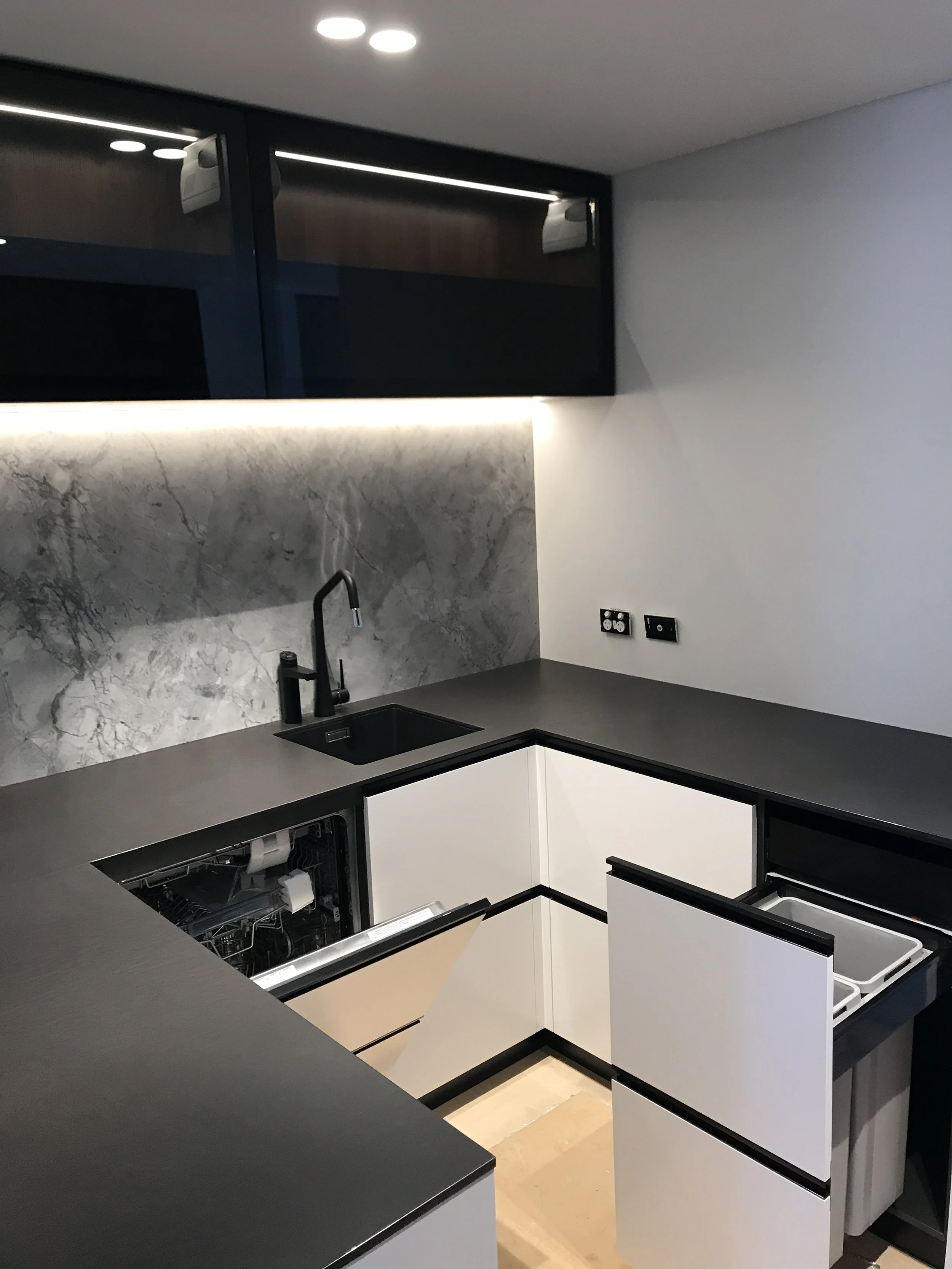 Modern kitchen with dark countertops, white cabinets, marble backsplash, and built-in dishwasher, with black hardware and a black faucet.