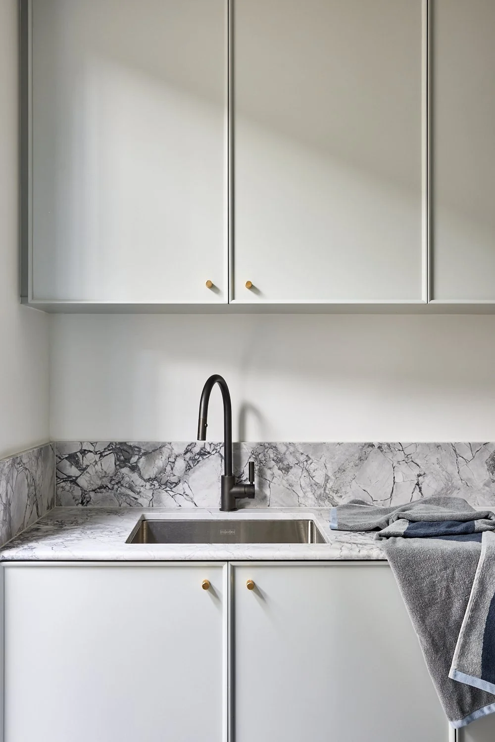 A minimalist kitchen with white cabinets, a marble countertop, and a black faucet over a stainless steel sink, with folded gray and dark gray towels on the right side.