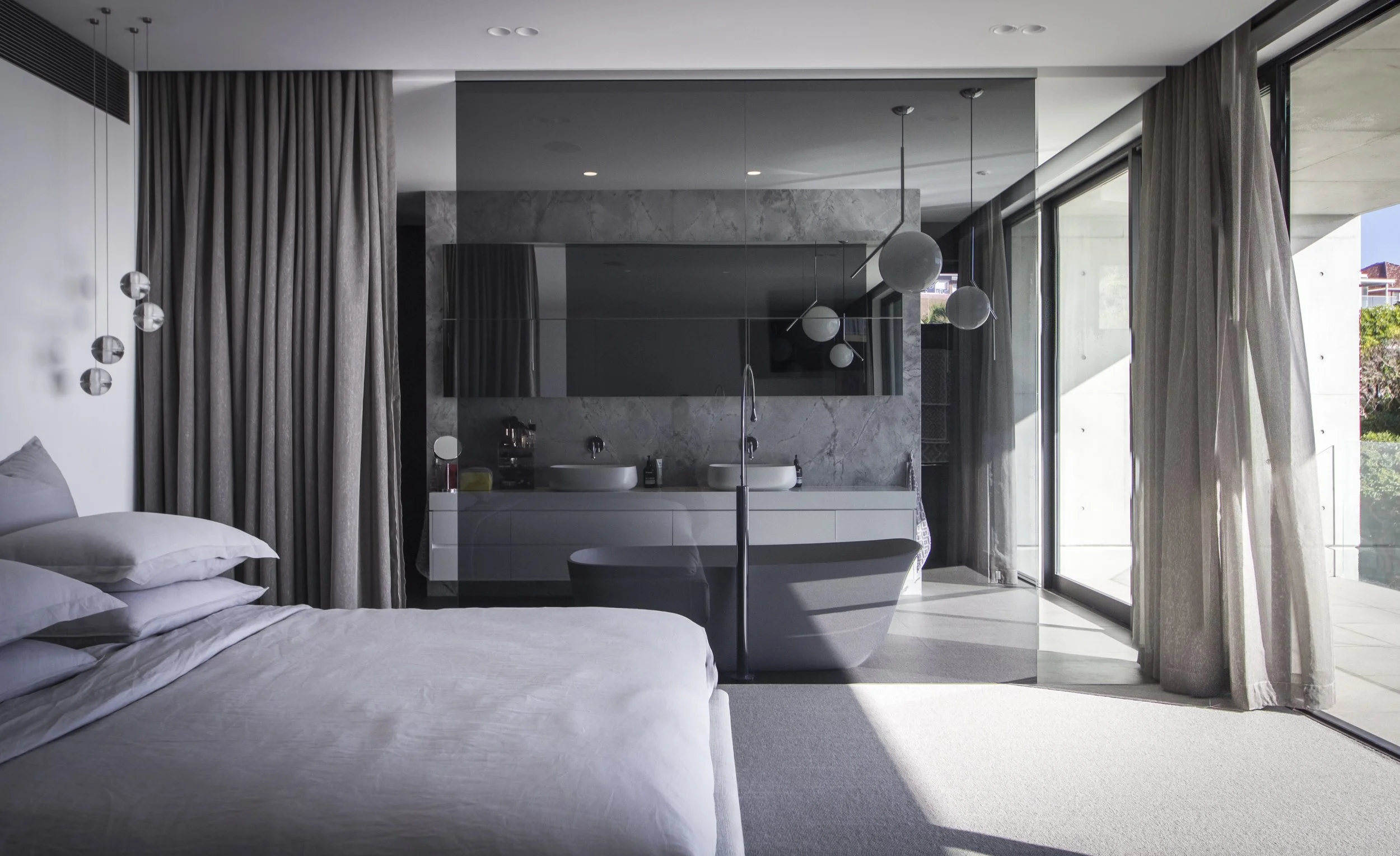 Modern bedroom with white bedding, large curtains, and a view of glass doors leading to a balcony, with a bathroom area visible behind a glass partition.