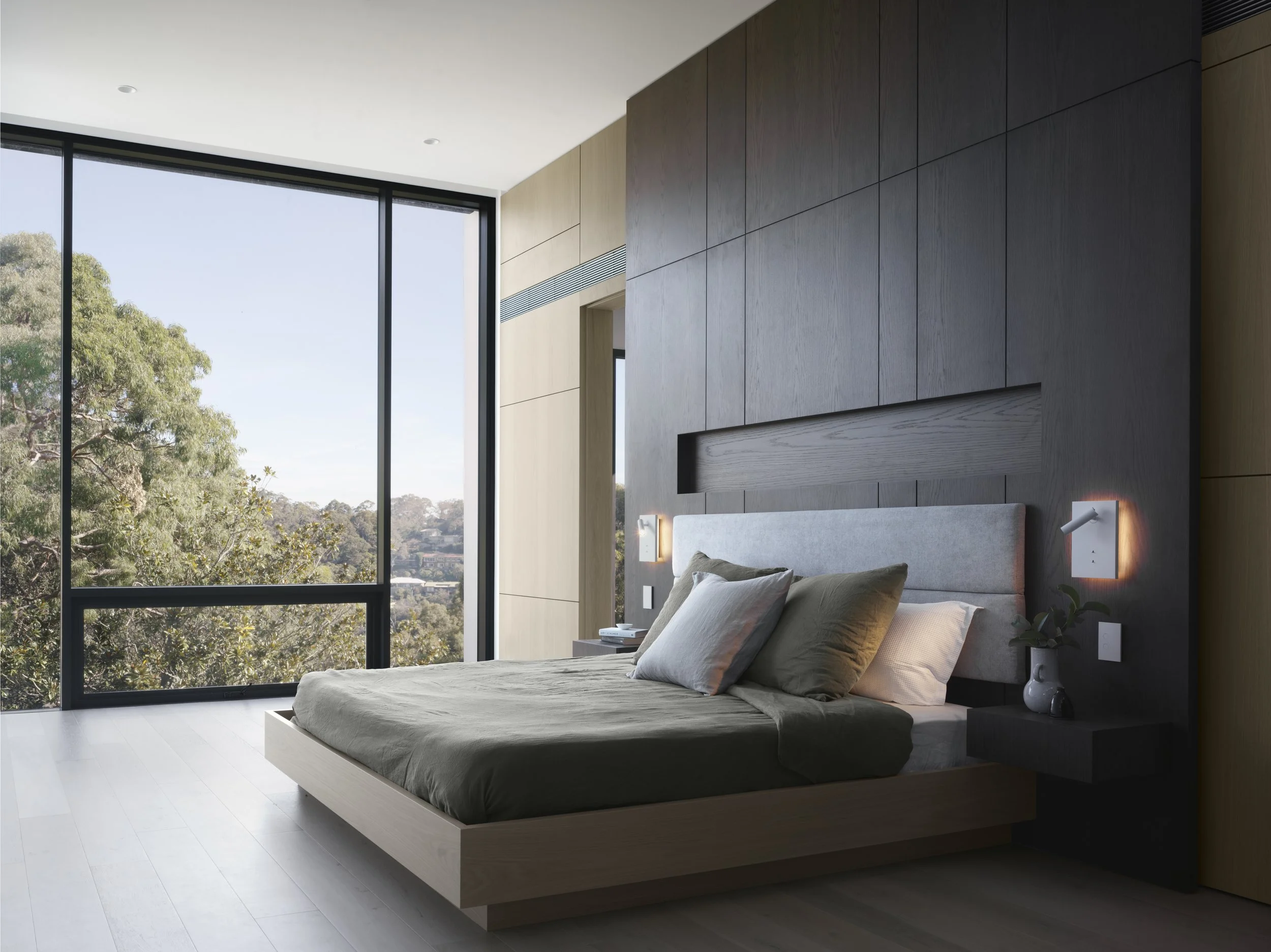 Modern bedroom with large floor-to-ceiling windows showing trees outside, a bed with beige bedding and gray pillows, and minimalistic furniture including a black bedside table with a white vase, in a bright and airy space.