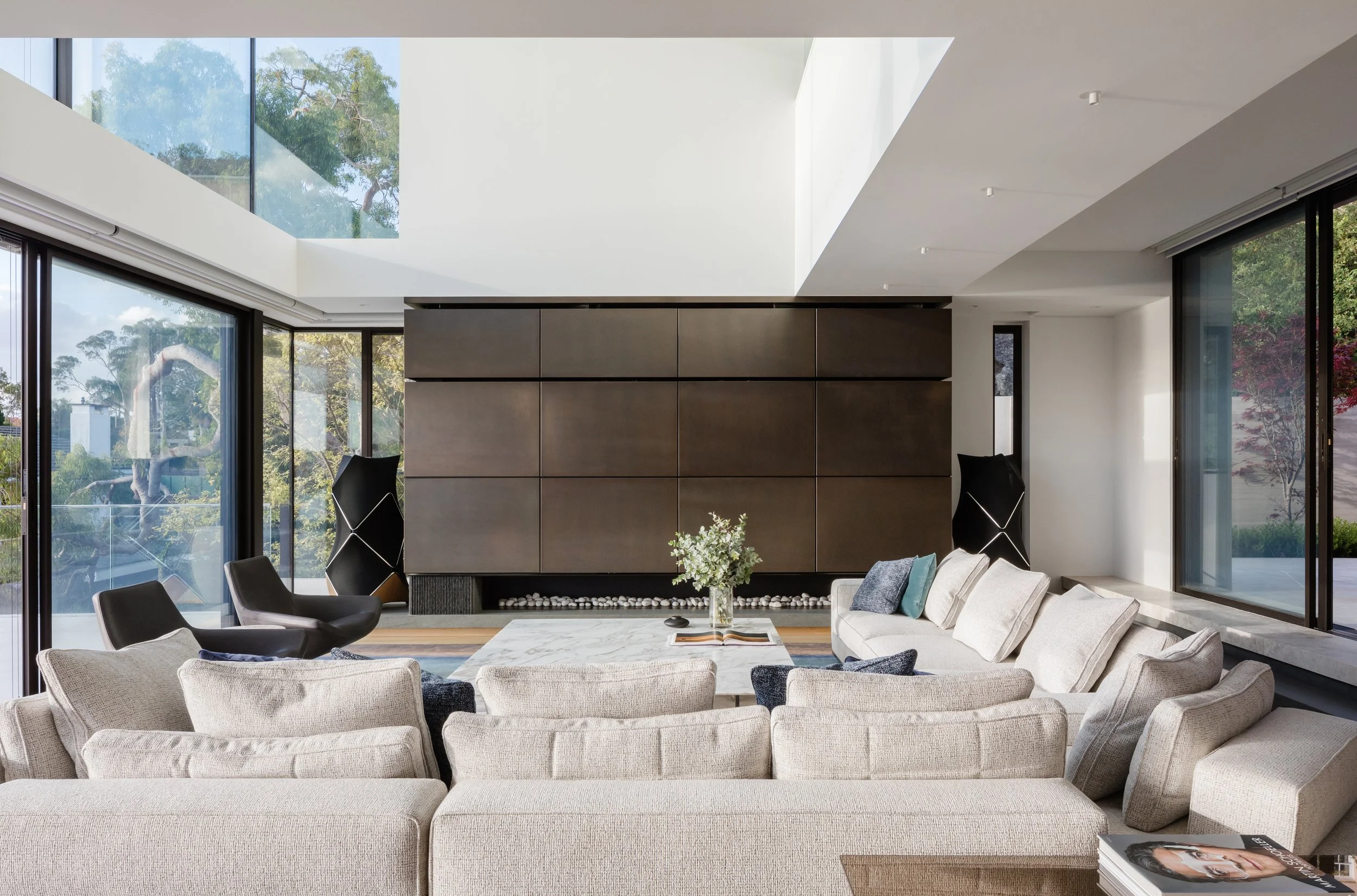 Modern living room with beige sectional sofa, black armchairs, a marble coffee table with a flower vase and an open book, large floor-to-ceiling windows, and a dark wood wall unit. Outside trees are visible through the windows.