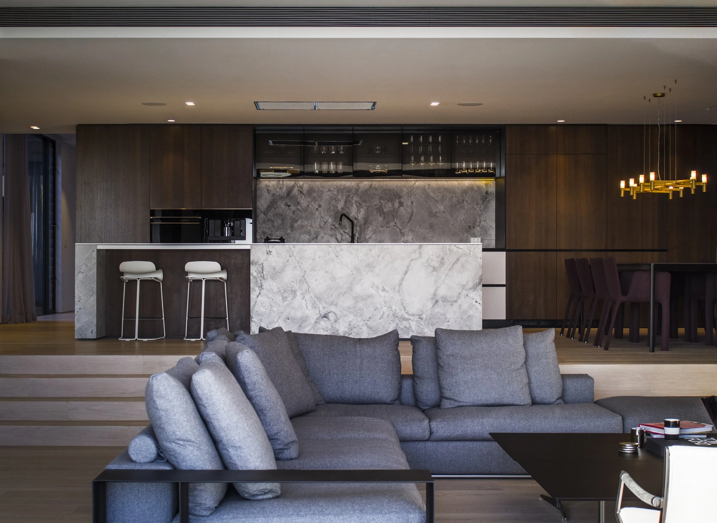 Modern living room featuring a gray sectional sofa with cylindrical cushions, a black coffee table, and a kitchen with a marble island and wood cabinetry. A large, gold chandelier hangs from the ceiling, and part of a dining area with dark chairs is 