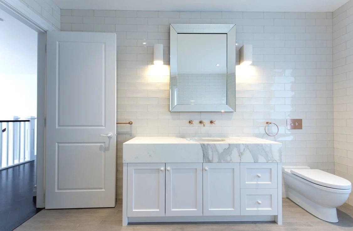 Modern bathroom with white subway tile walls, a marble vanity with cabinet storage, a rectangular mirror, wall-mounted light fixtures, and a wall-mounted toilet.