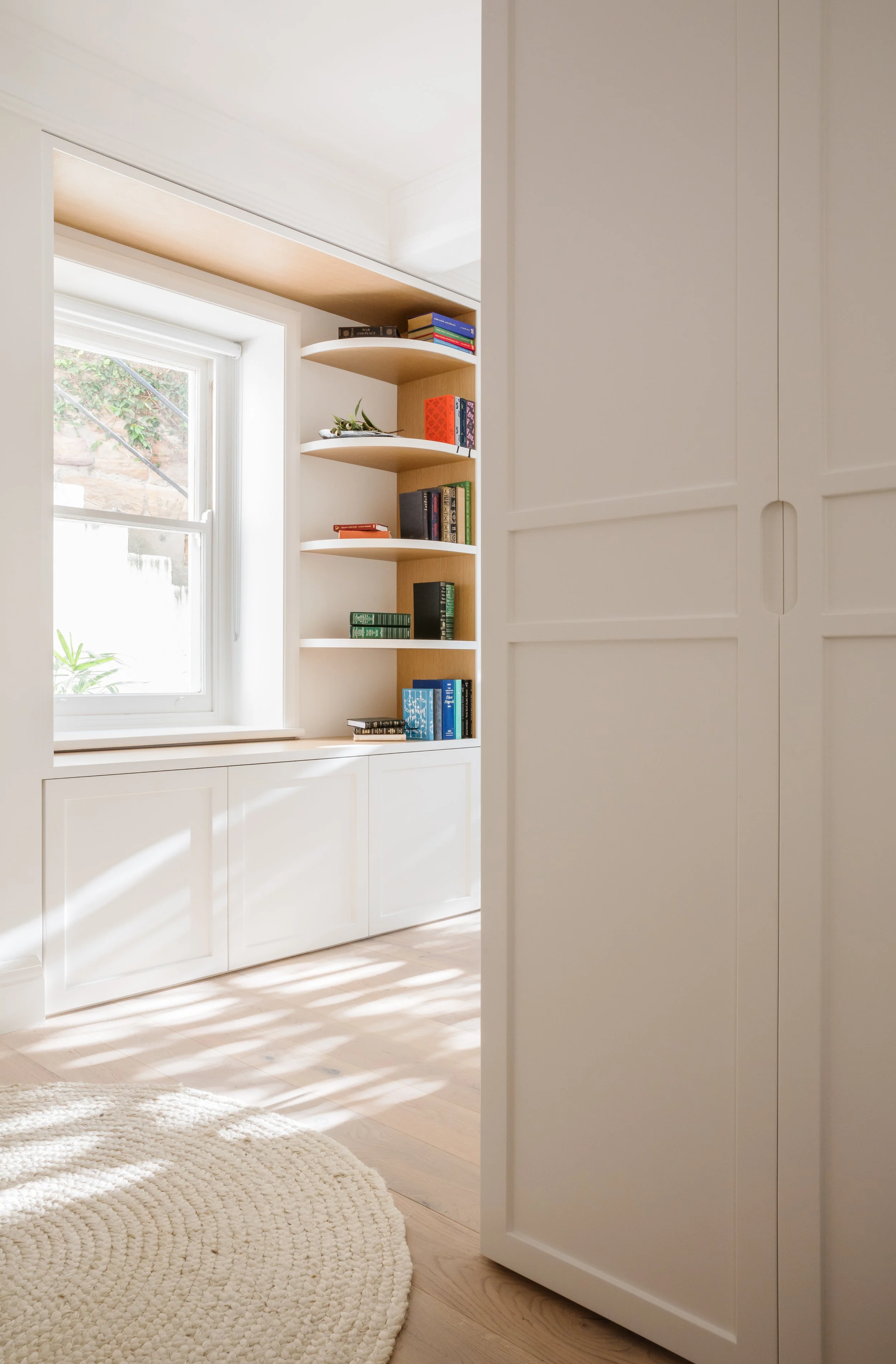 A cozy room corner with sunlight streaming through a window, a white built-in cabinet with paneled doors, a curved shelving unit filled with books and a small plant, a cream woven rug, and light wooden flooring.