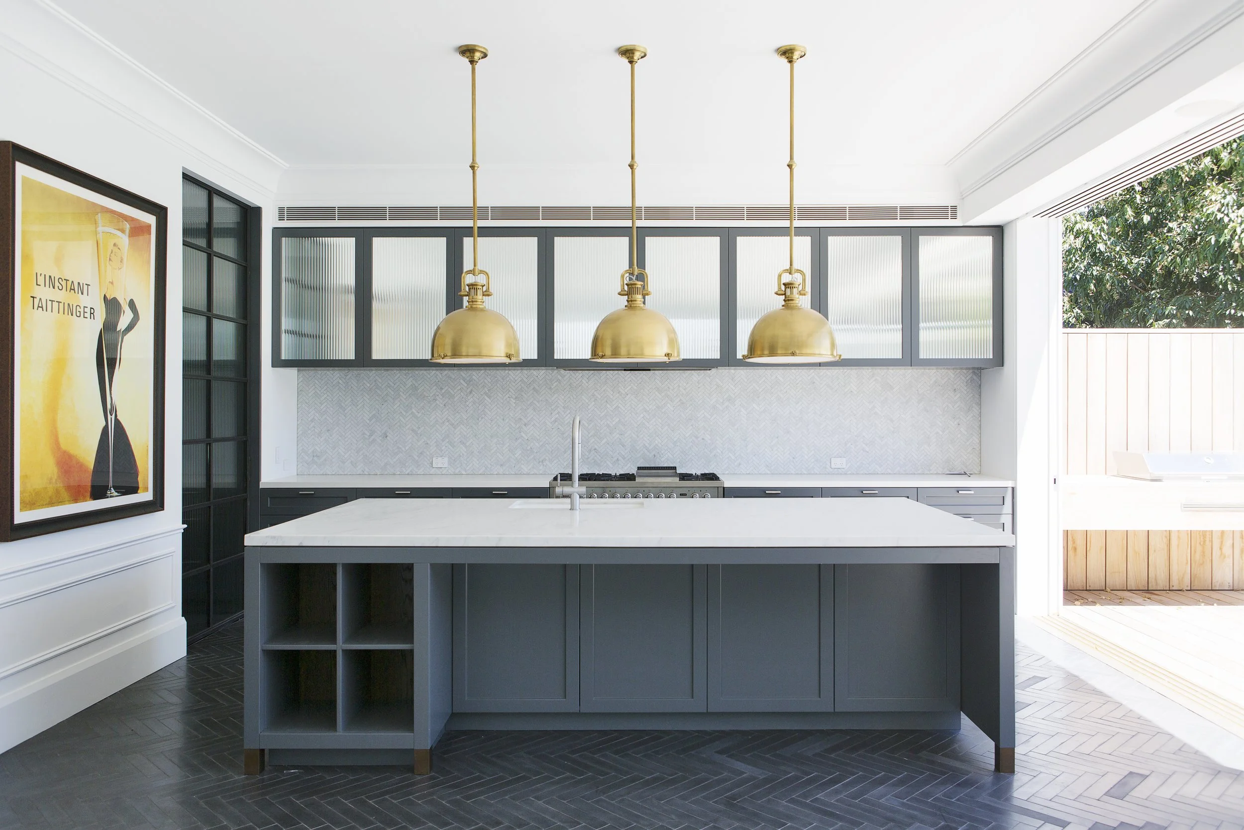 Modern kitchen with navy blue cabinetry, white marble island, and three gold pendant lights. Artwork on the wall portraying a woman in a black dress and yellow background. Large window with blinds, leading to an outdoor space with greenery.