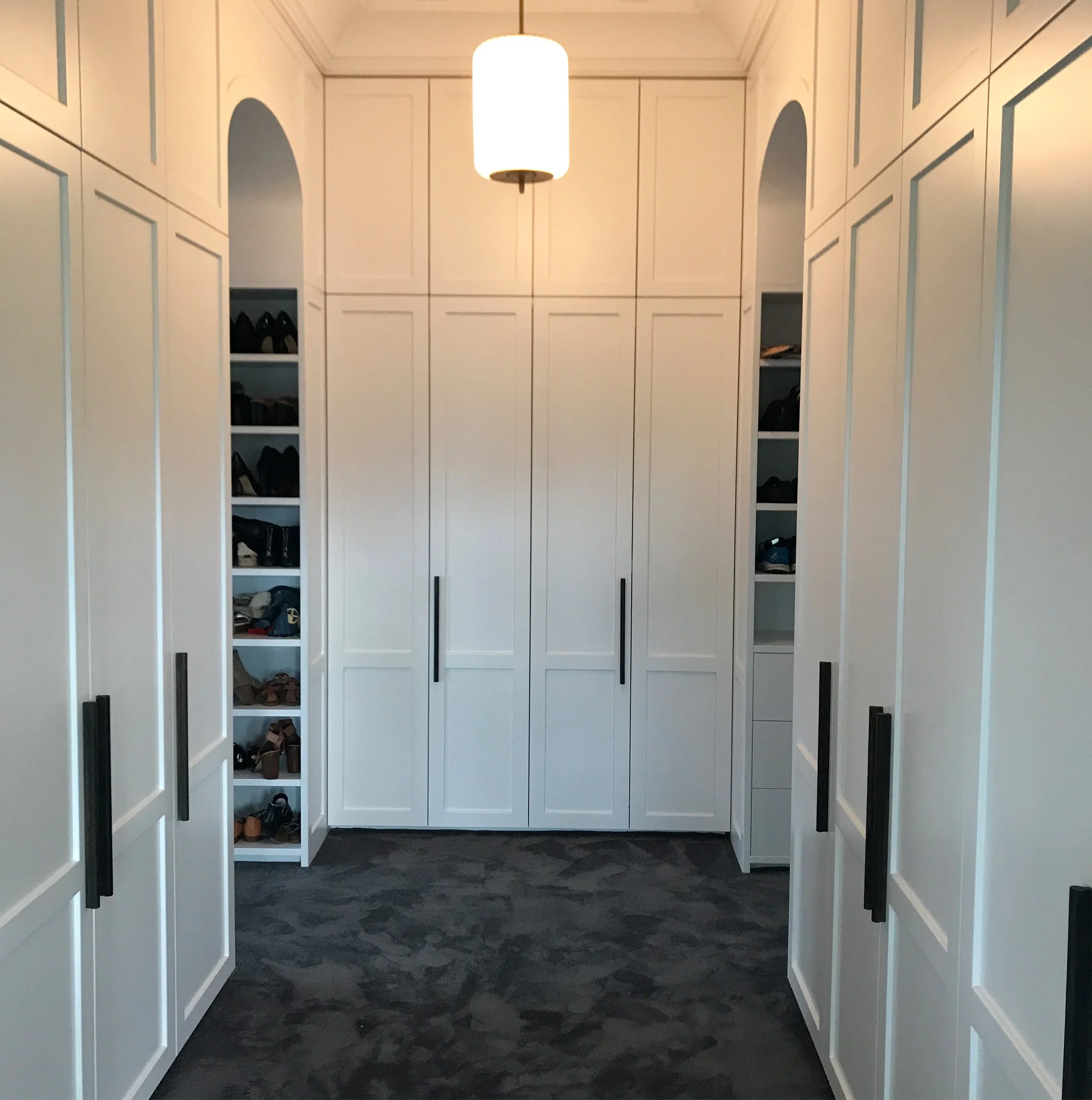 A walk-in closet with white built-in shelves and cabinets. Shoes and other items are stored on open shelves on both sides. A large white cabinet with closed doors is at the end of the closet. The floor has a dark, patterned carpet, and a white ceilin