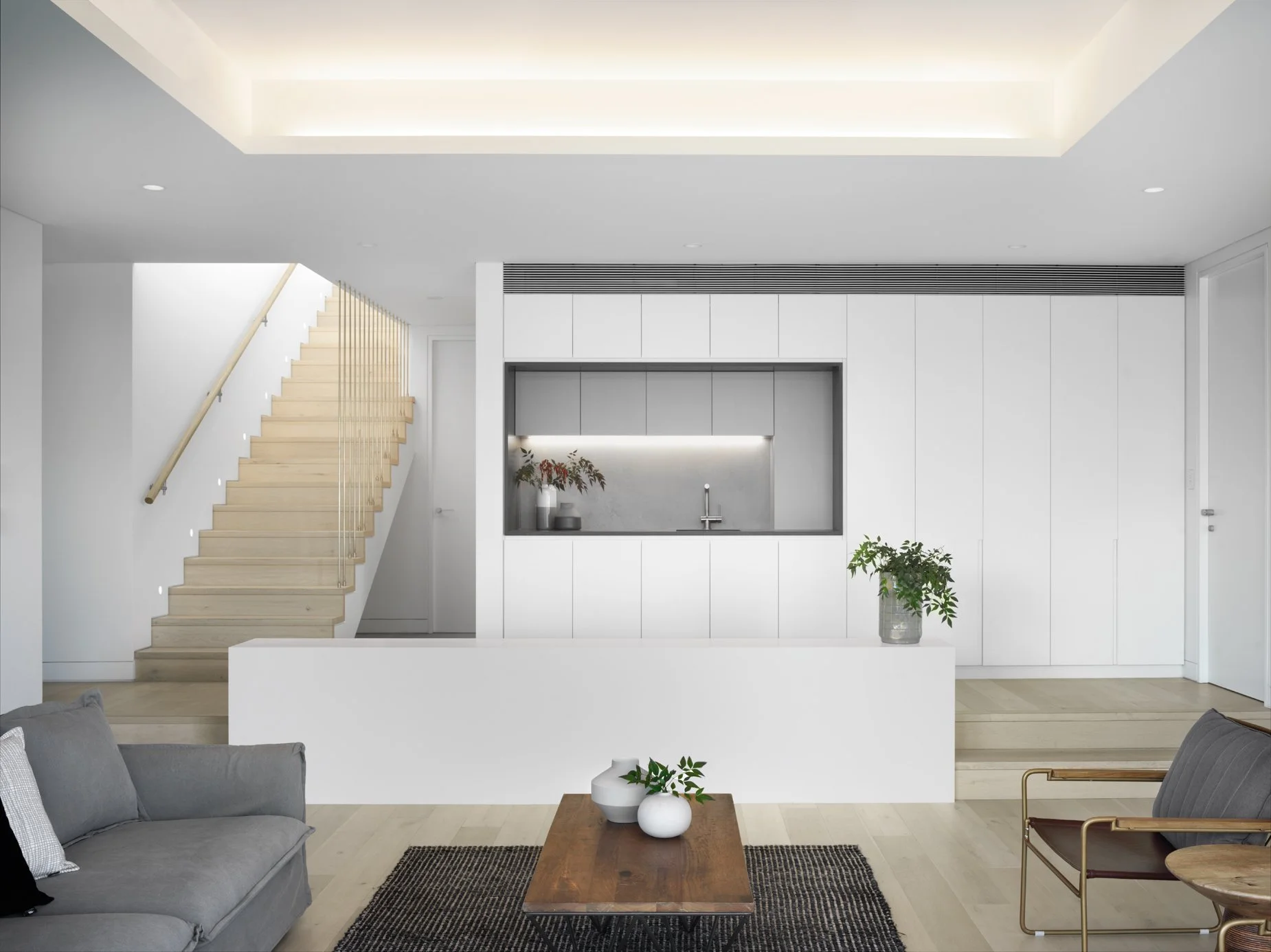 Modern minimalist living room with a gray sofa, wooden coffee table, and a black rug. White walls with a staircase on the left, and a built-in kitchen area with a sink, green and red plants, and white cabinetry in the background.