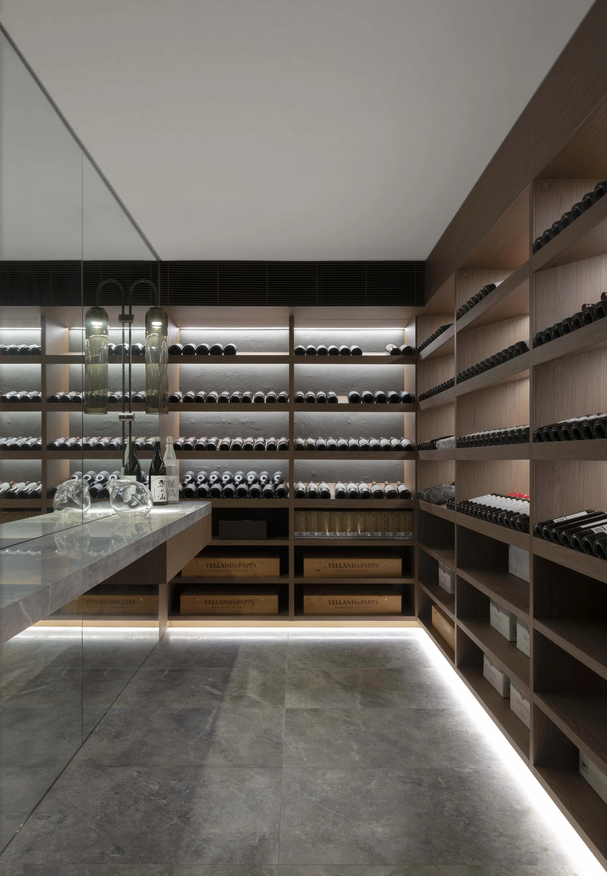 A wine cellar with wooden shelves filled with bottles, a marble-topped counter with glass decanters, and a glass wall reflecting the room.