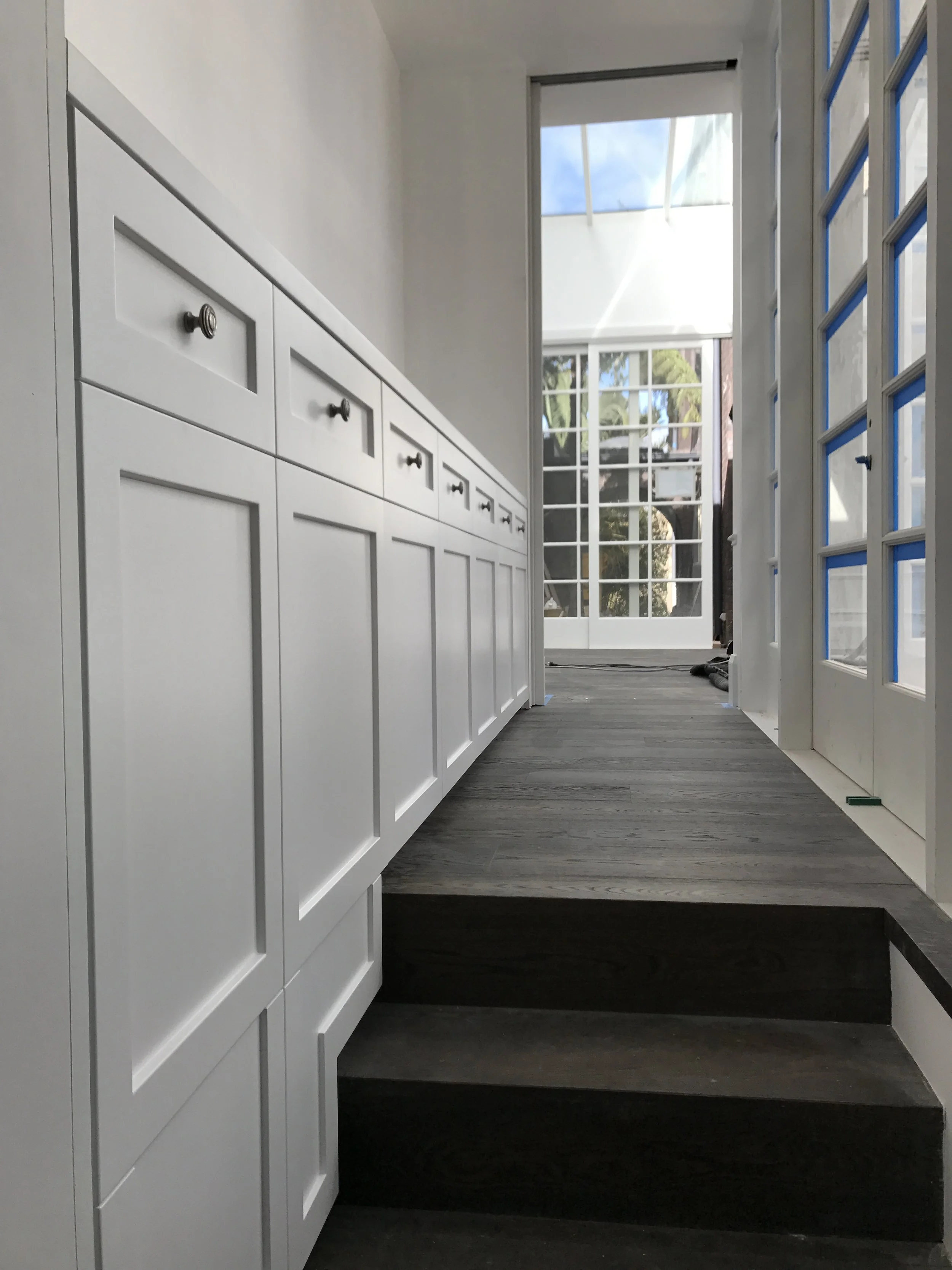 Interior view of a modern room with white cabinets on the left, a staircase at the bottom, and a set of glass-paneled doors at the end leading to an outdoor area with trees and a bright sky.