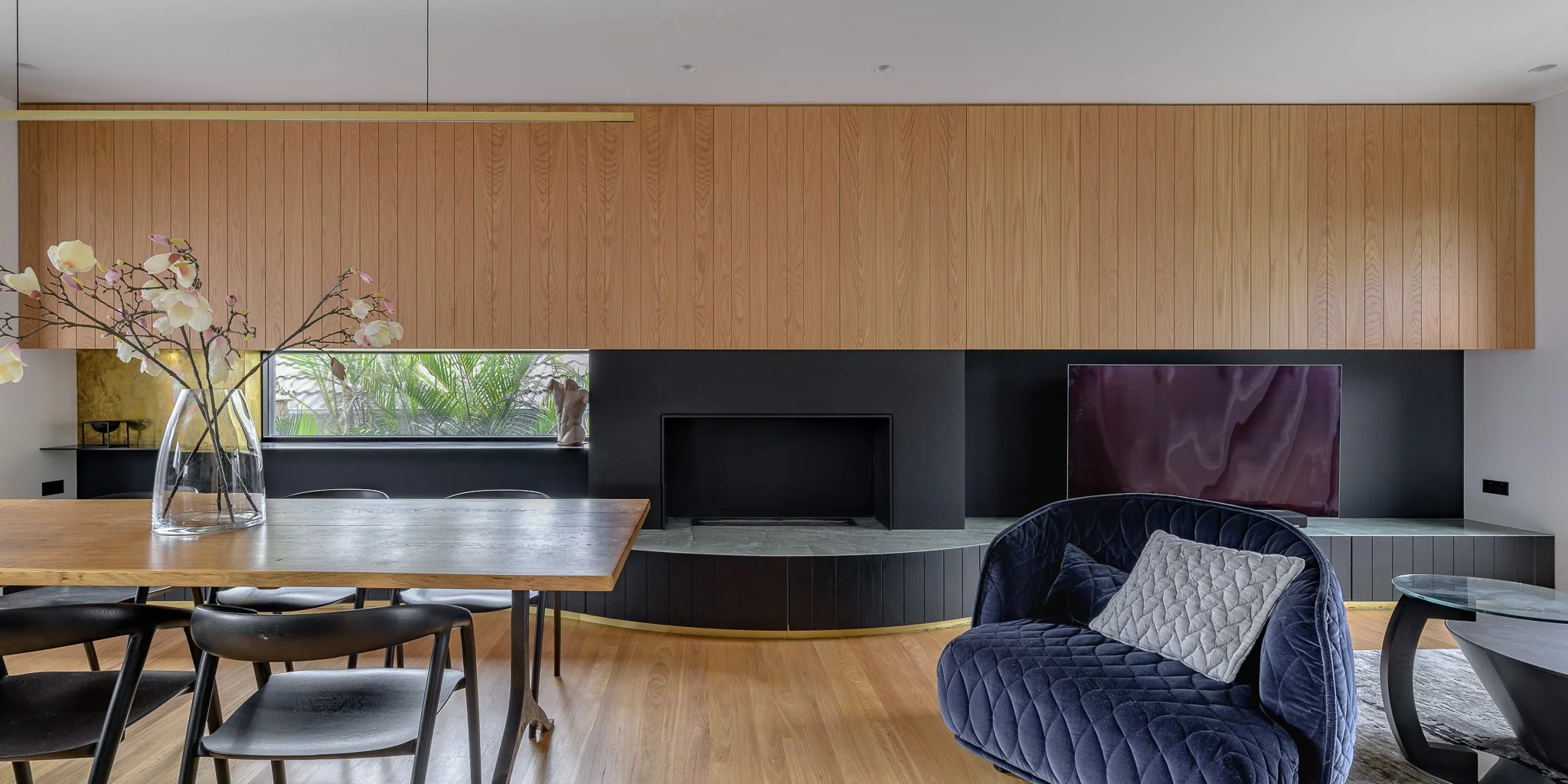 Modern living room with a wooden ceiling, black fireplace, a TV, a blue velvet armchair with a pillow, a wooden dining table with black chairs, and a glass vase with white flowers.