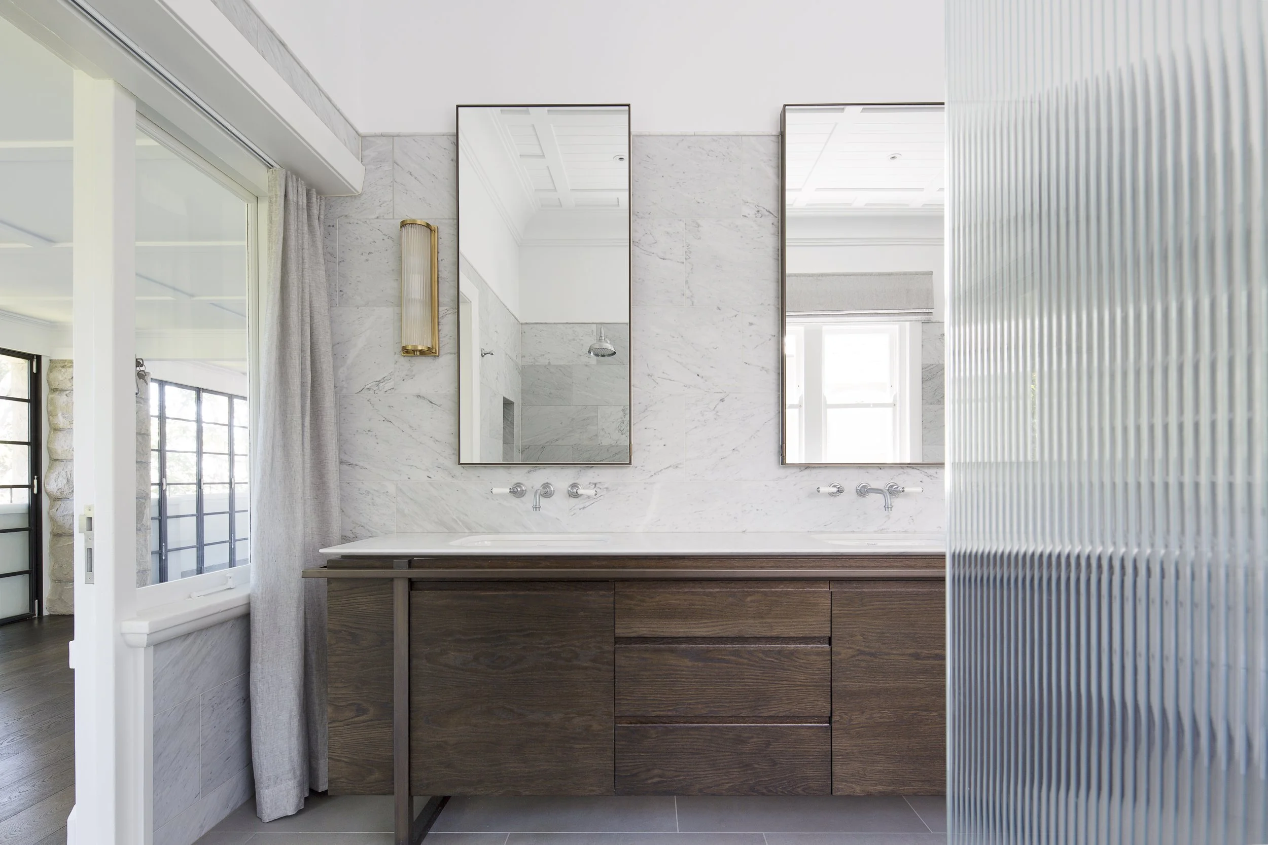 Modern bathroom with white marble wall tiles, a large rectangular mirror above a dark wooden vanity, a brass wall light, a window with cream curtains, and a textured glass partition.