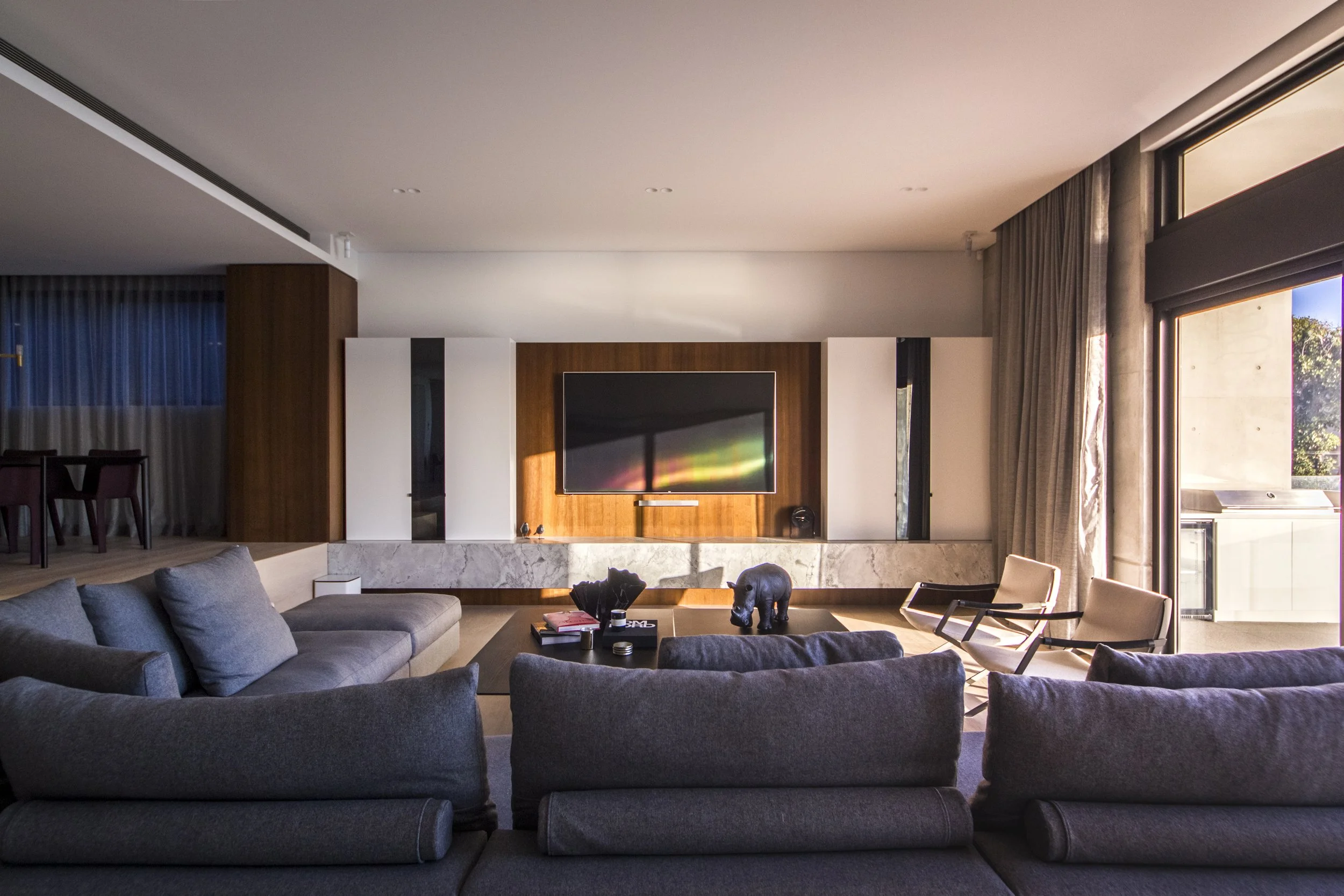 Modern living room with gray sectional sofa, black elephant sculpture on coffee table, wall-mounted TV, large windows with curtains, and exposed concrete and wood paneling.
