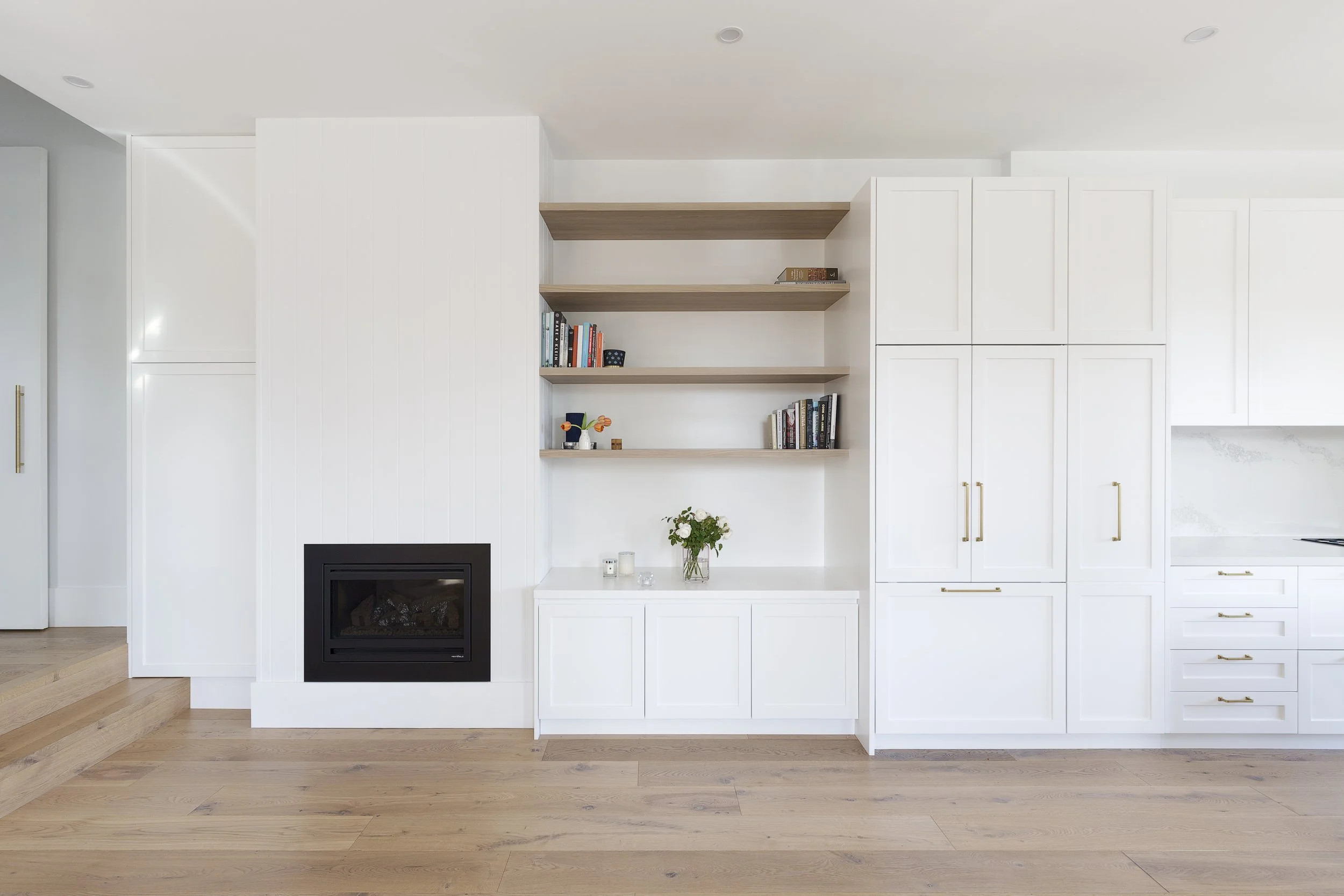 Interior of a modern kitchen featuring white cabinets, open wooden shelves with books and decorative items, a black fireplace, and wood flooring.