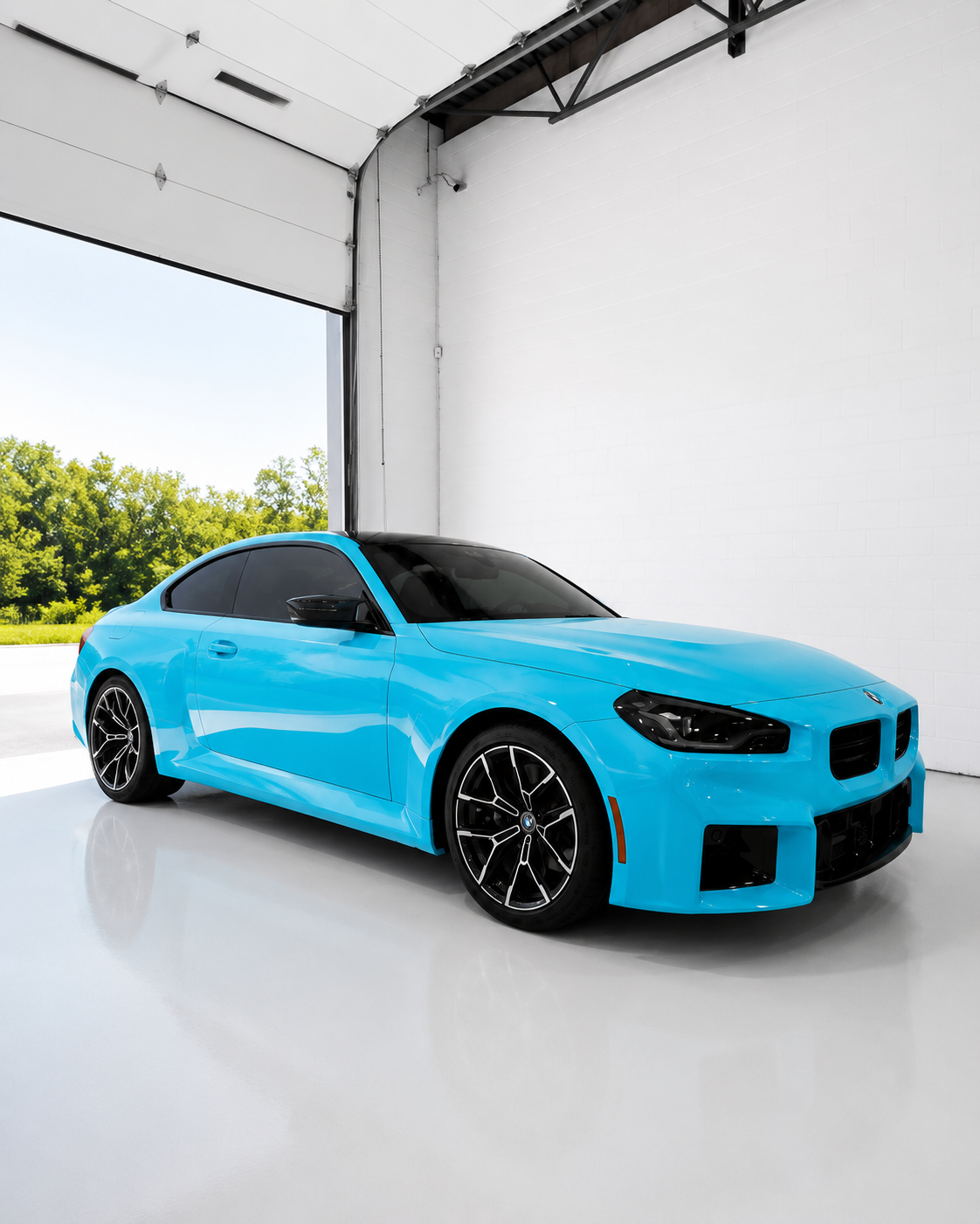 A blue BMW sports car parked inside a bright, industrial-style garage with a white floor and walls, open garage door revealing green trees outside.