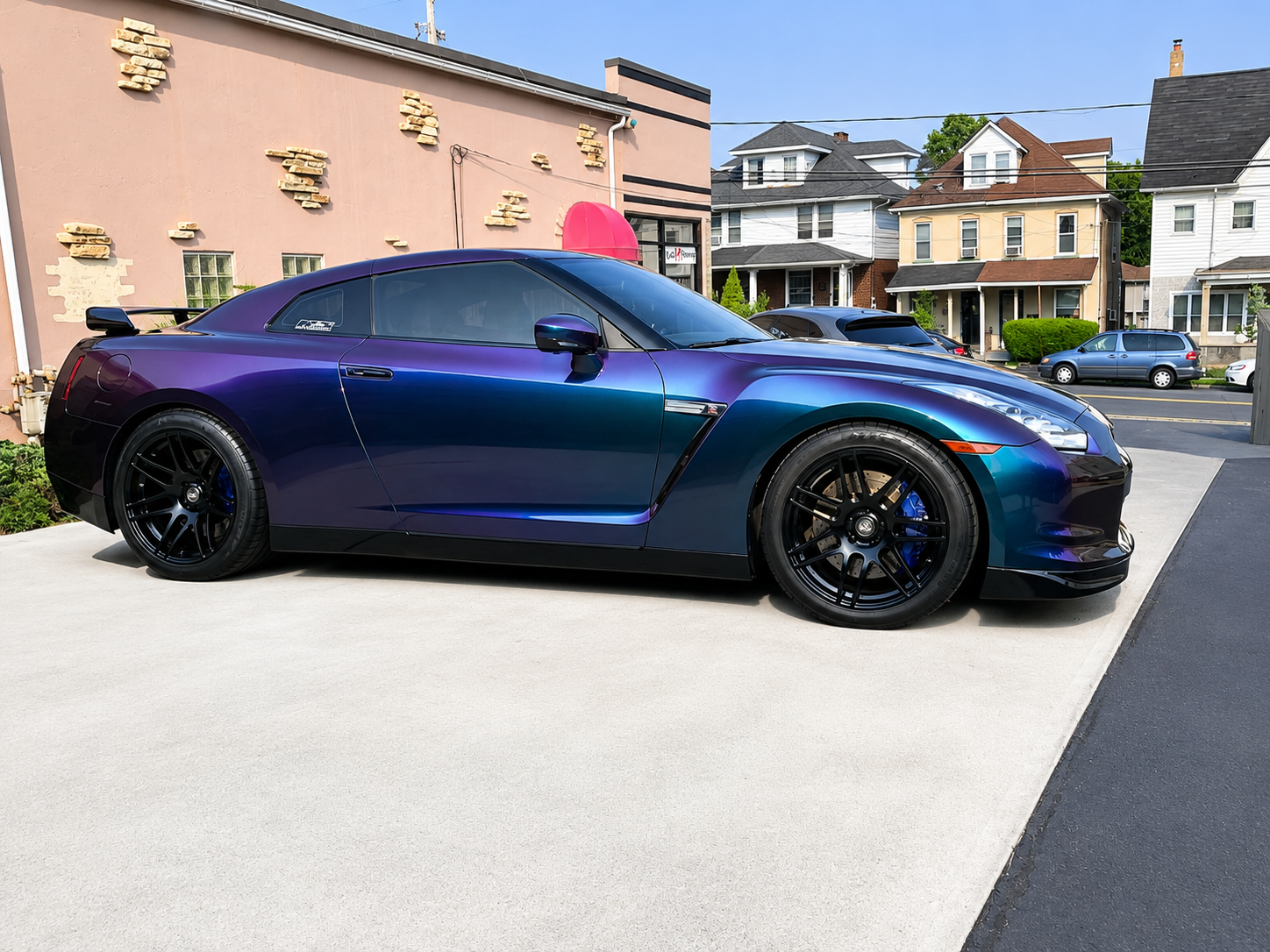 A purple and blue sports car parked on a light-colored concrete driveway in a residential neighborhood with multicolored houses and other parked cars.